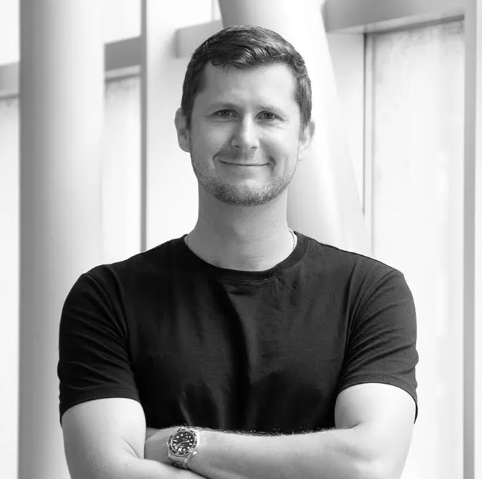 Smiling man with short hair and light facial hair wearing a black t-shirt and a wristwatch, standing with arms crossed in front of a window.