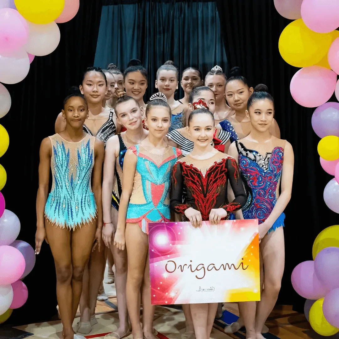 Group of young rhythmic gymnasts in colorful leotards standing together, with two girls in front holding a sign that says 'Origami', surrounded by pink, yellow, and white balloons.
