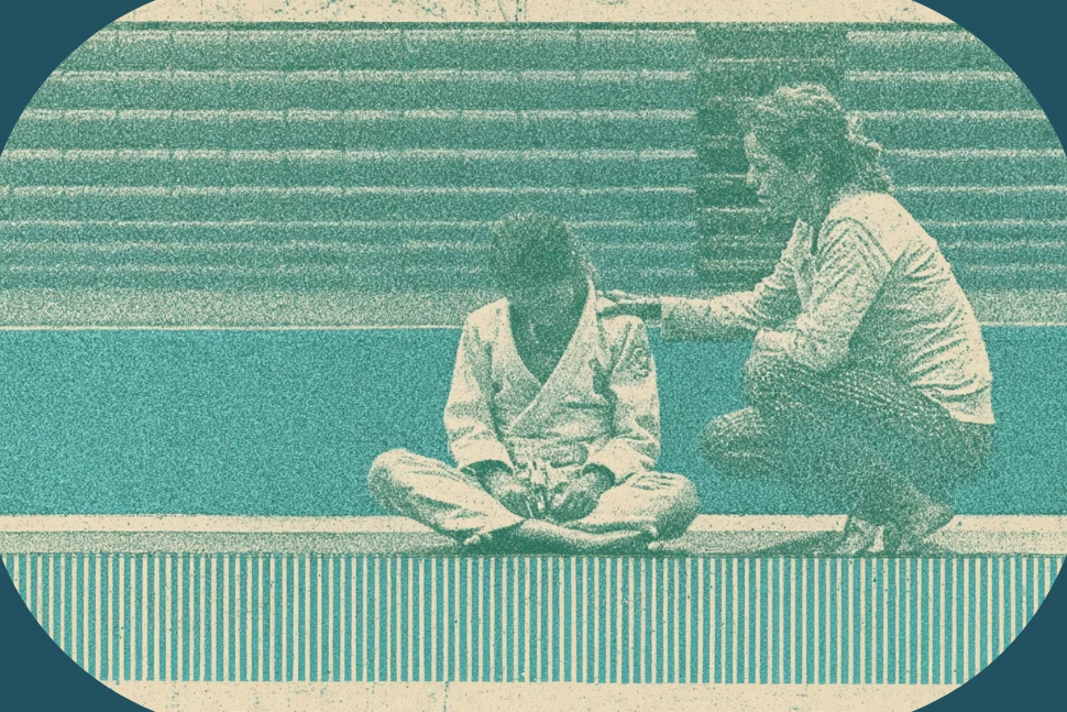 A coach crouches beside a young jiu-jitsu competitor sitting on the edge of a tournament mat after a lost match