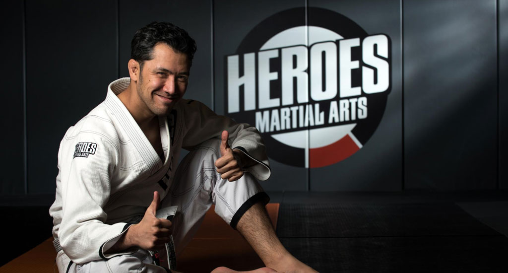 Alan Gumby Marques in a white gi giving a thumbs up in front of the Heroes Martial Arts logo