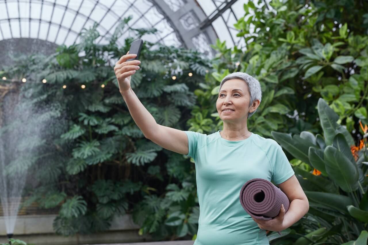 Woman with short gray hair holds a rolled yoga mat and takes a selfie in a greenhouse full of tropical plants
