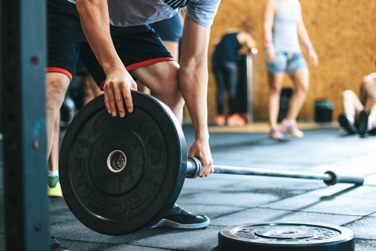 Person loads a bumper plate onto a barbell on a rubber gym floor while other athletes train in the background