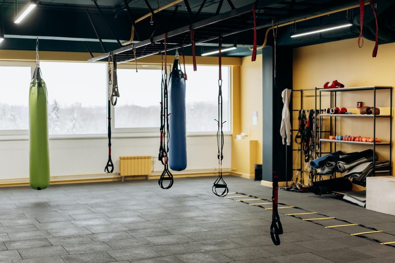 Empty gym with hanging punching bags, suspension straps, rubber flooring, and shelves of mats and gear