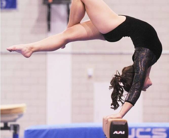 Gymnast in a black leotard performing a handstand with split legs on a balance beam in a gymnastics gym