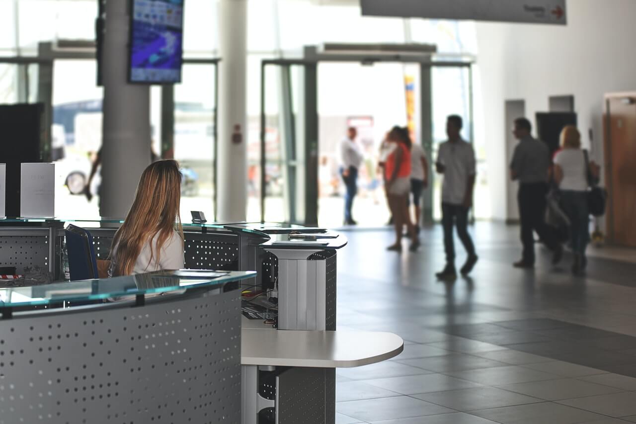 Staff member seated at a front reception desk in a busy lobby as people walk past the entrance