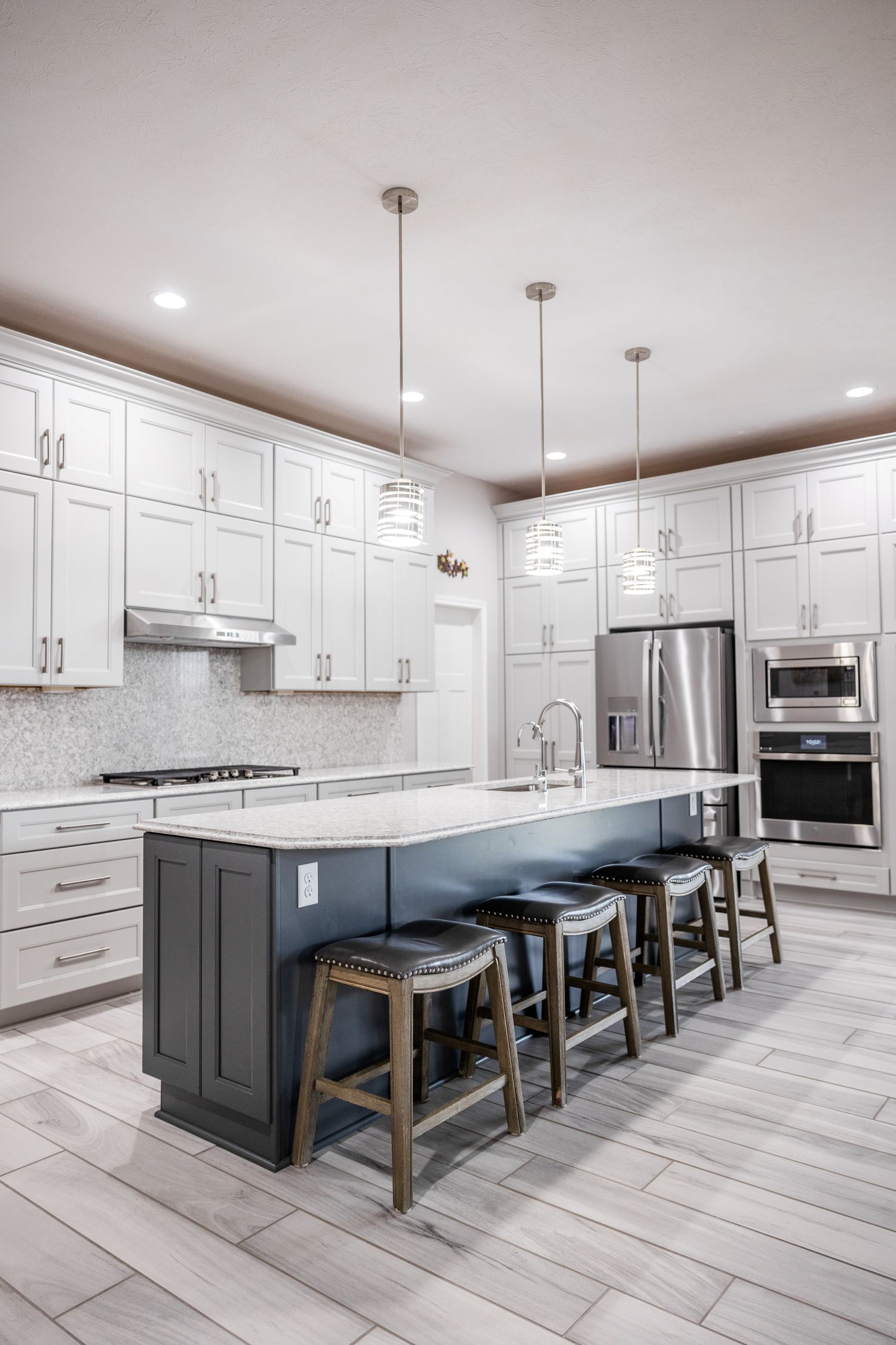 Modern kitchen with a large dark island topped with white marble, four wooden stools, white cabinets, stainless steel appliances, and three pendant lights.