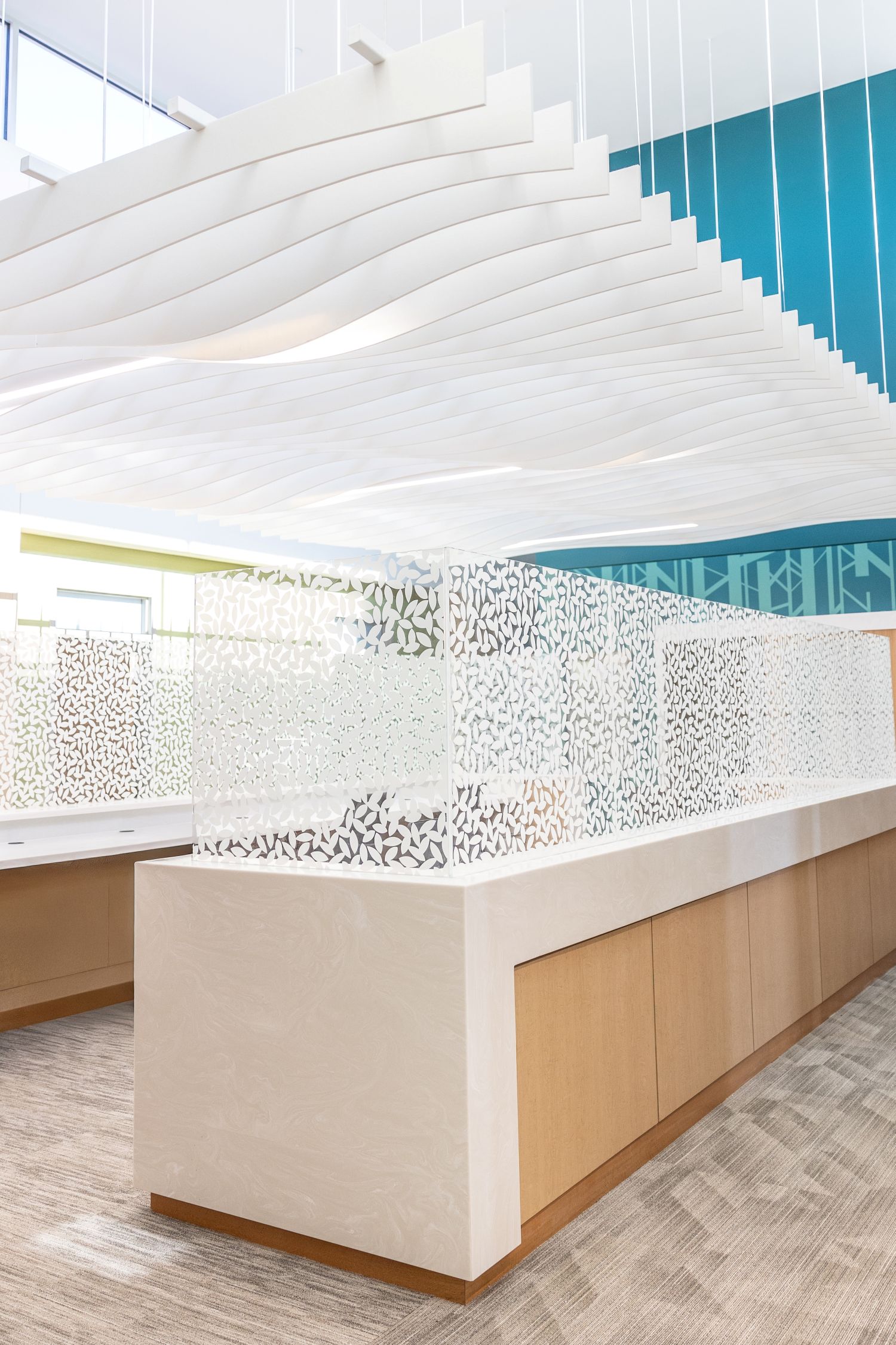 Modern office interior with white wave-like ceiling panels, frosted glass partitions with leaf patterns, wooden cabinets, and carpeted floor.