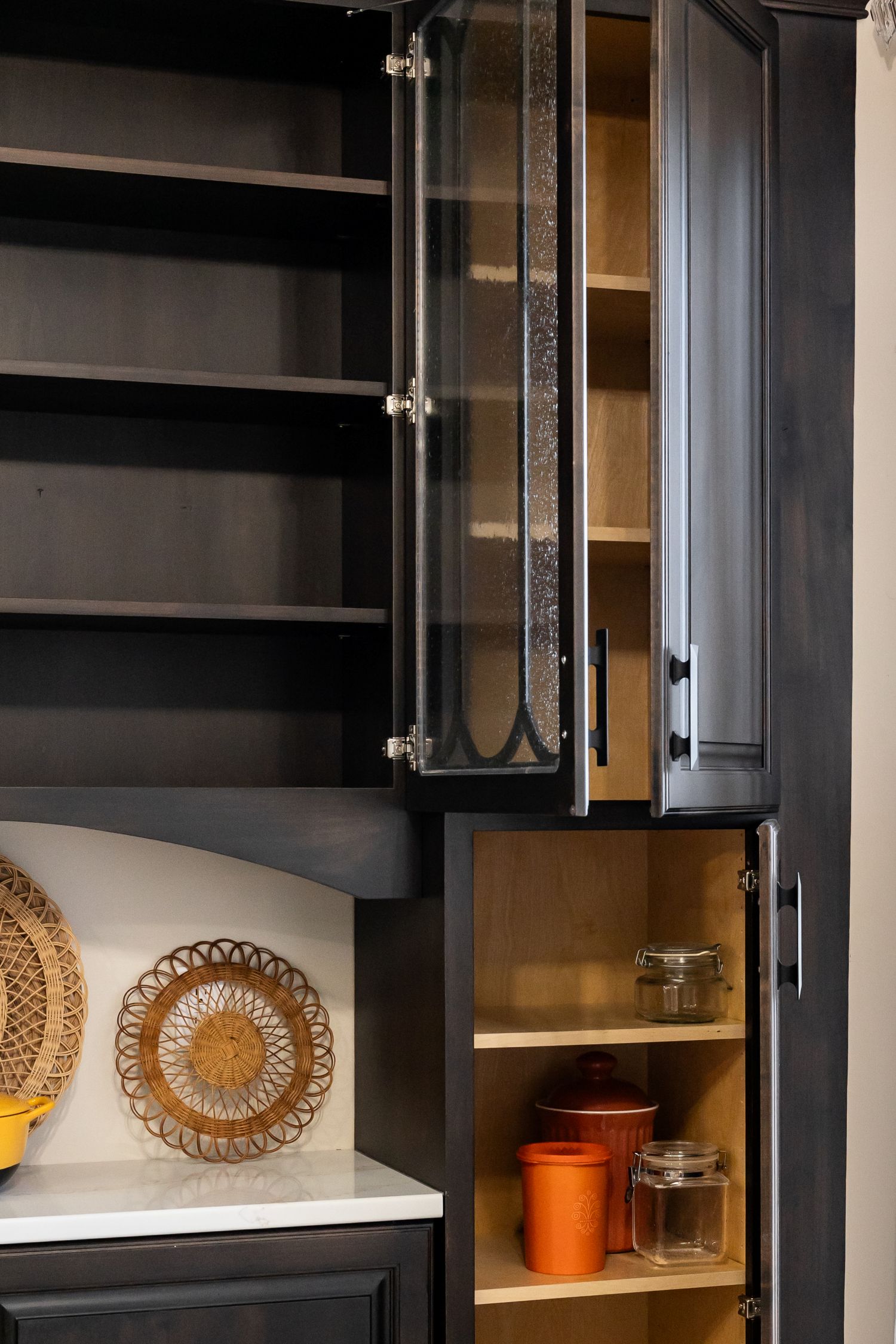 Dark wooden kitchen cabinets with glass-paneled doors and shelves holding ceramic and glass jars, next to a marble countertop.