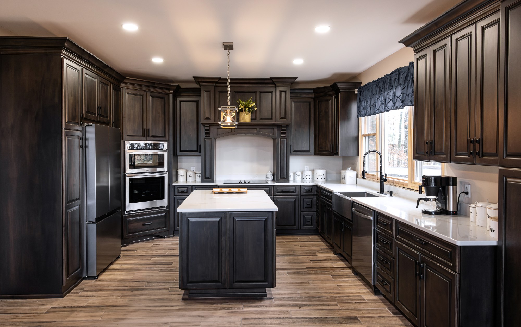 Modern kitchen with dark wood cabinetry, white countertops, stainless steel appliances, and a central island under recessed lighting.