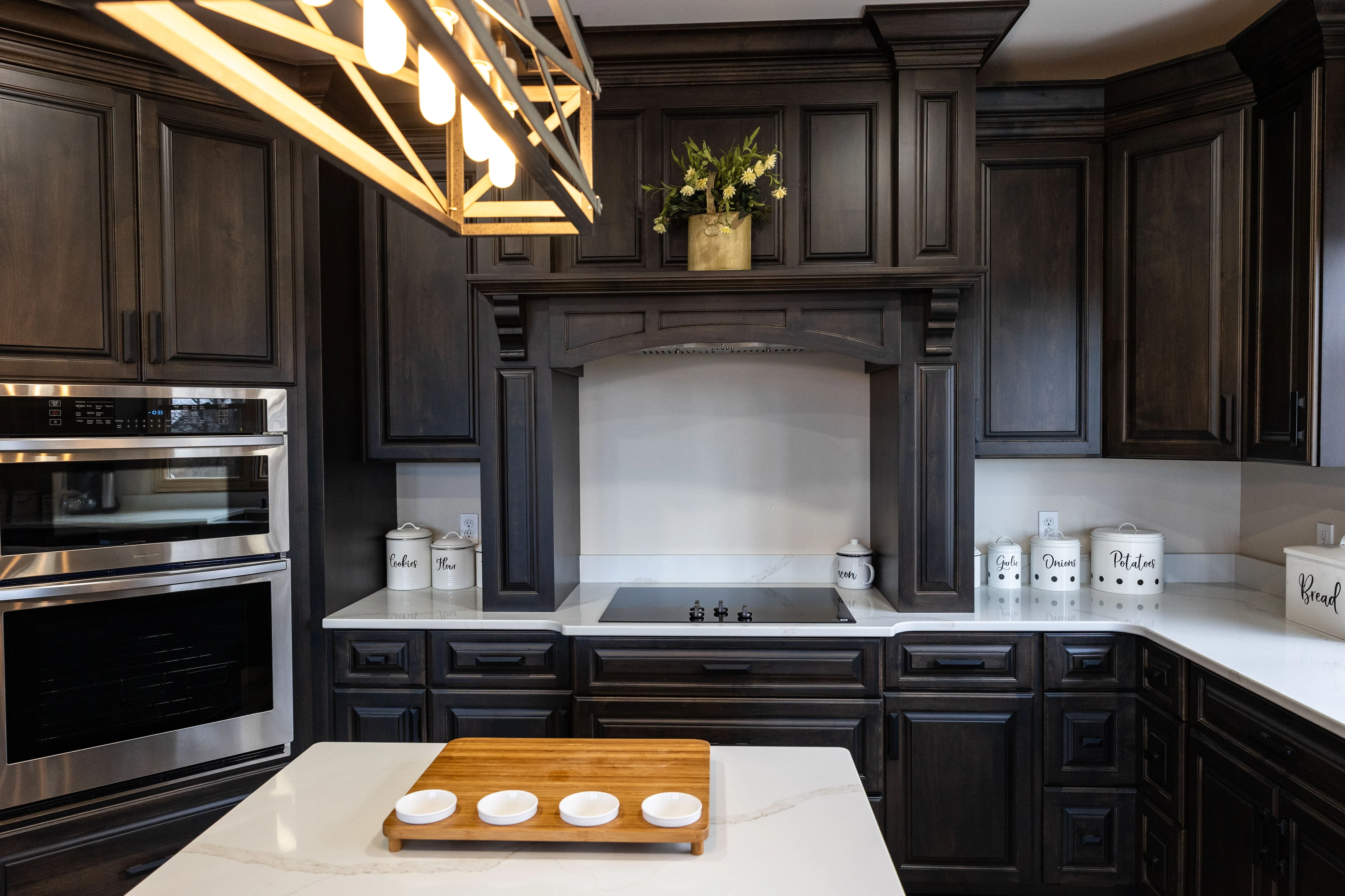 Modern kitchen with dark wood cabinets, white countertops, stainless steel double oven, cooktop under a decorative hood, and labeled white canisters on the counters.