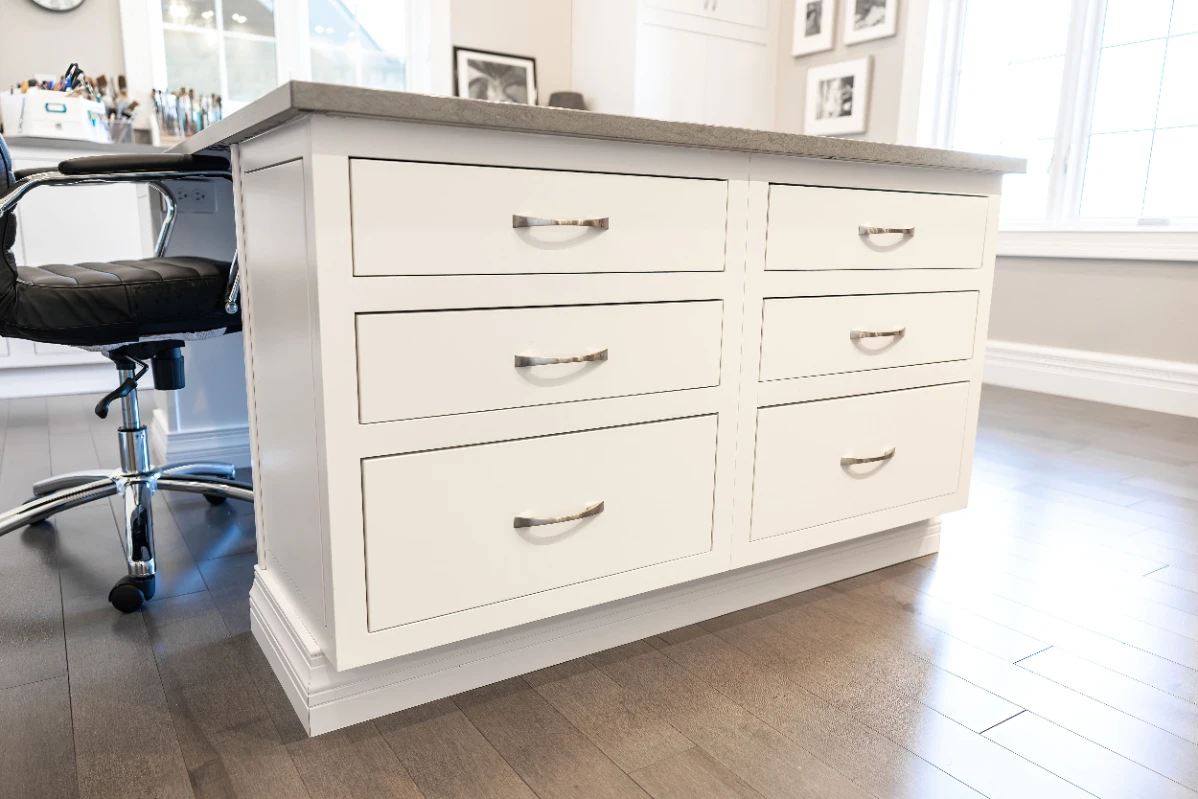 White wooden cabinet with six drawers and chrome handles beside a black office chair on dark wood floor in a bright room.