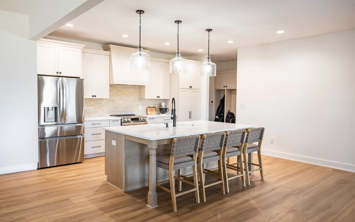 Modern kitchen with a large island, four woven-back chairs, stainless steel refrigerator, white cabinets, and three pendant lights.