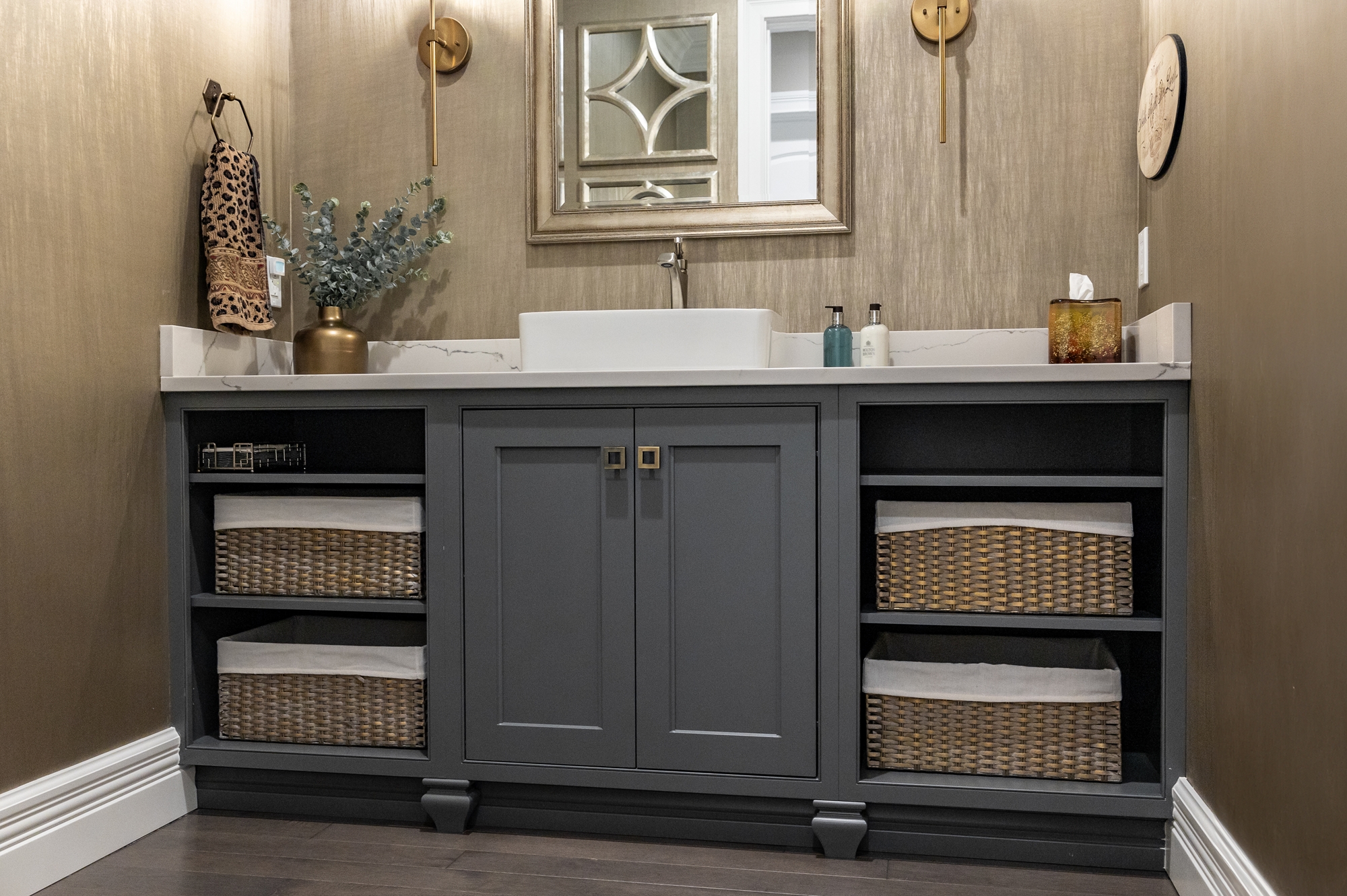 Modern bathroom vanity with gray cabinets, white vessel sink, wicker baskets, and decorative items on marble countertop.