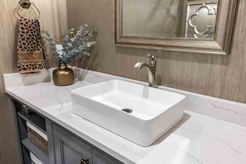 Modern bathroom vanity with a rectangular white vessel sink, a silver faucet, decorative mirror, and a gold vase with green foliage on the countertop.