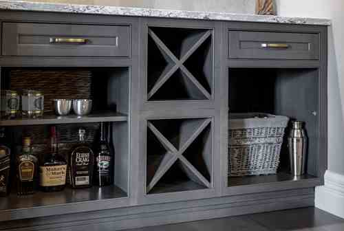 Gray wooden bar cabinet with open shelves holding liquor bottles, a metal shaker, a wicker basket, and X-shaped wine racks under a marble countertop.