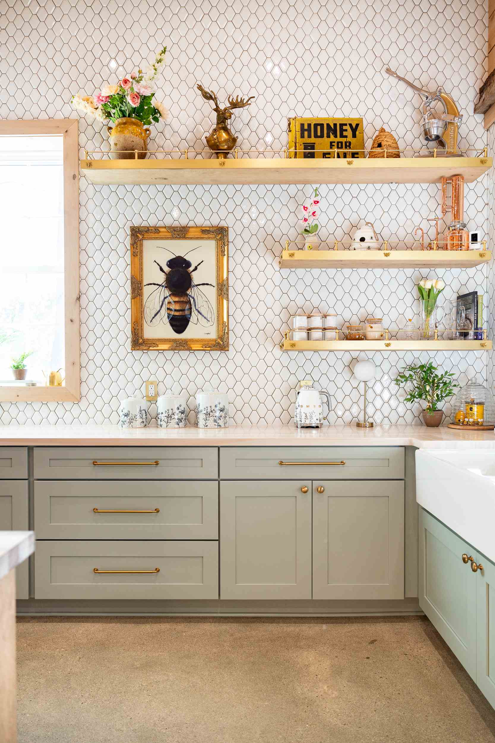 Bright kitchen corner with light gray cabinets, honeycomb tile backsplash, wooden shelves holding plants, jars, and a framed bee illustration.