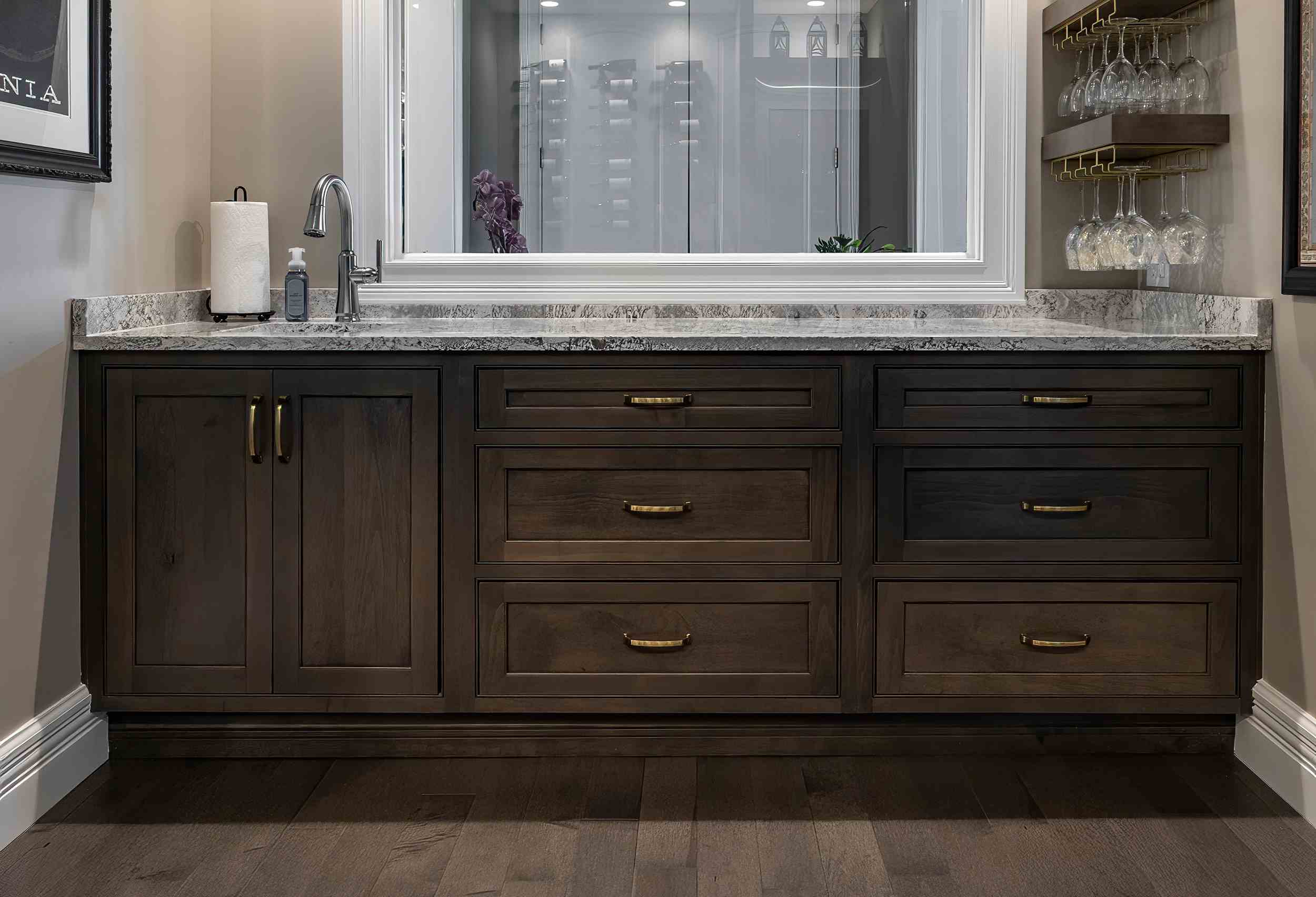 Dark wood bar cabinets with brass handles under a grey marble countertop with a small sink, faucet, paper towel and soap dispenser.