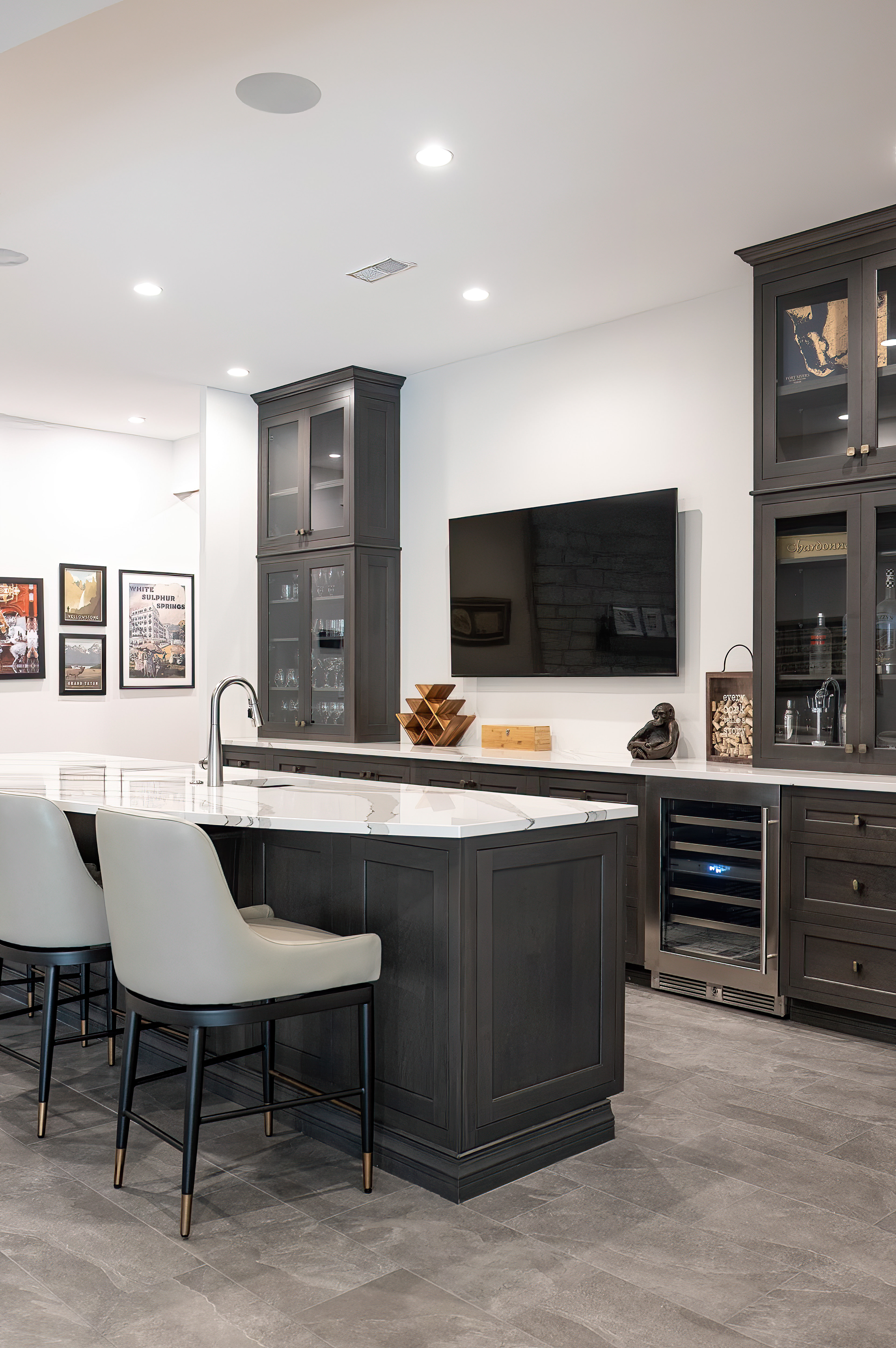 Modern kitchen with white marble countertop island, gray upholstered bar stools, dark wood cabinets, wall-mounted flat-screen TV, and wine cooler.