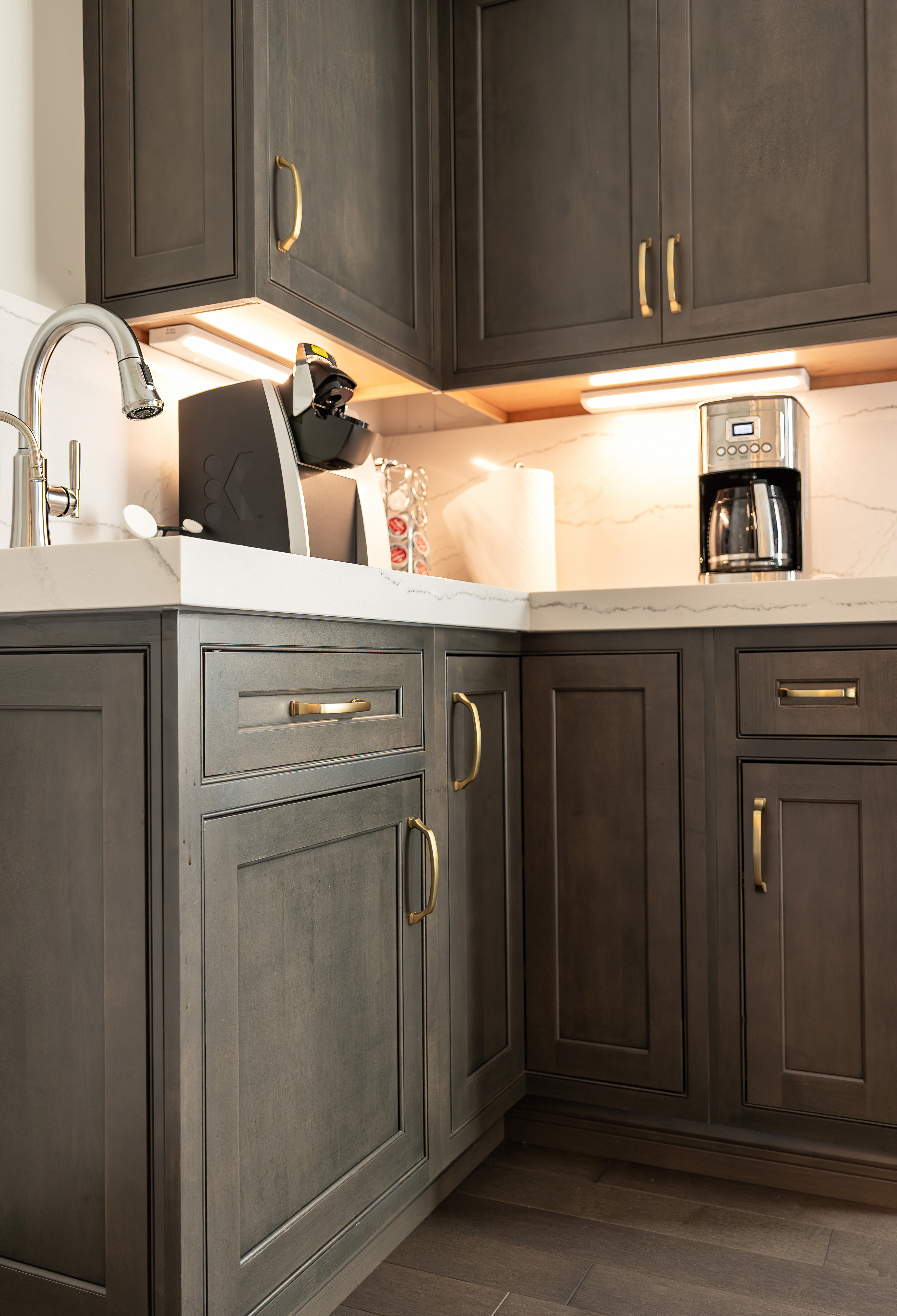 Modern kitchen corner with dark wood cabinets, brass handles, white marble countertops, coffee makers, and a stainless steel faucet.