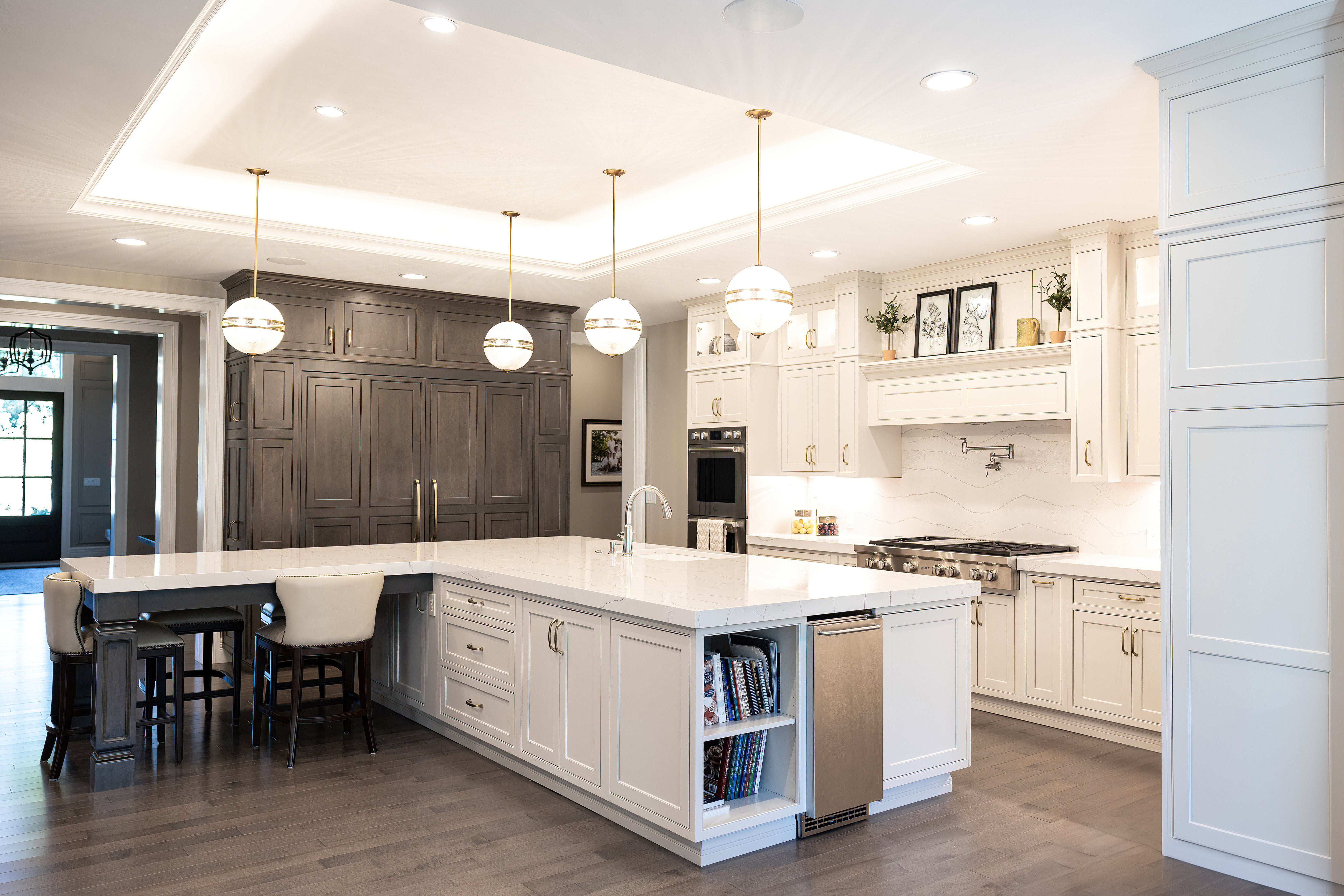 Bright modern kitchen with white cabinetry, large marble island with built-in sink and seating, pendant lights, and dark wood floor.