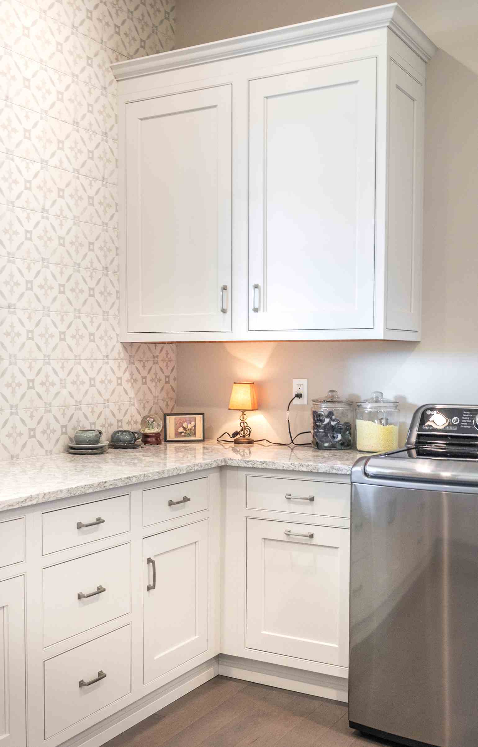 Laundry room corner with white cabinets, granite countertop, small table lamp, glass jars with laundry pods and powder, and a stainless steel washing machine.