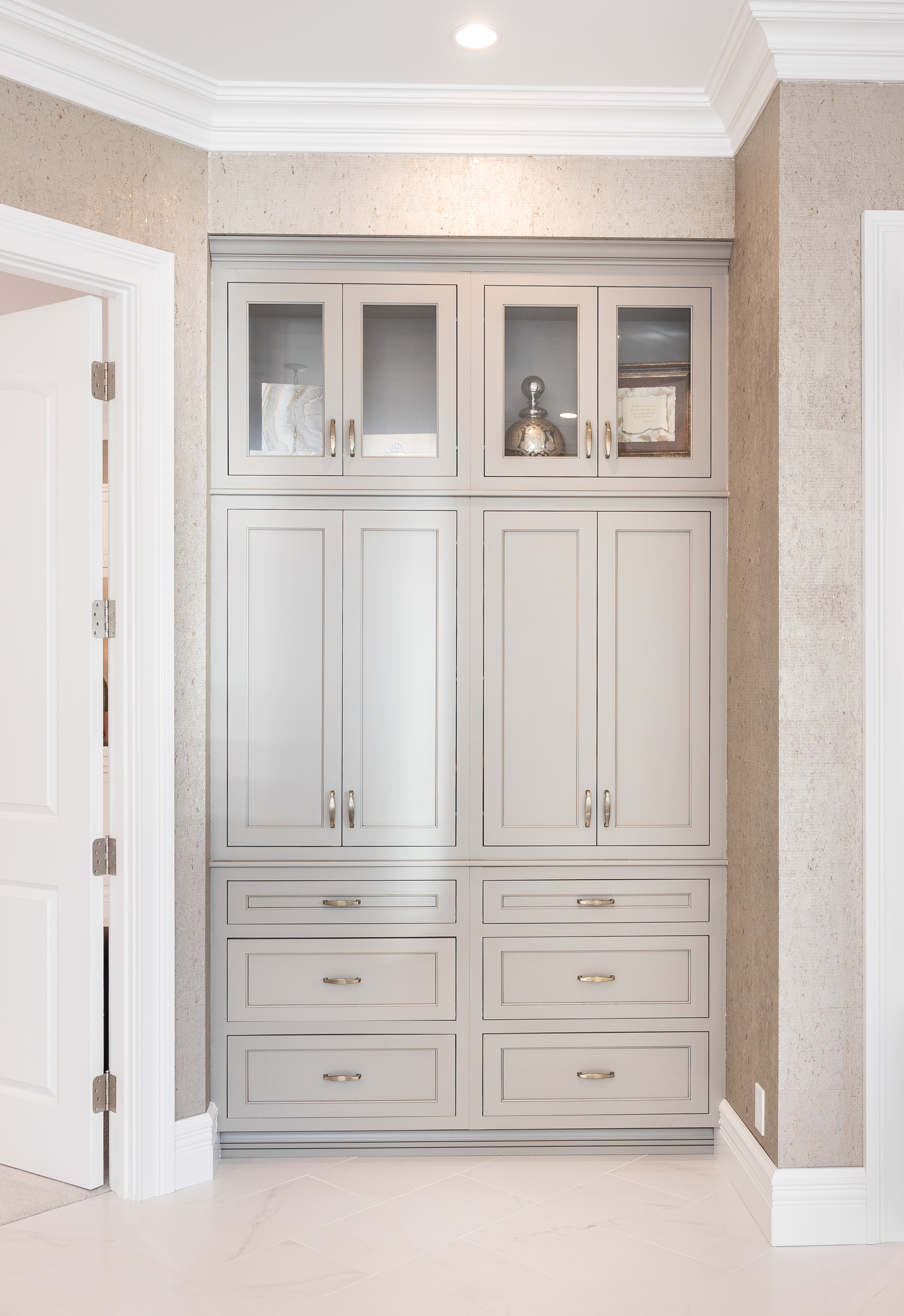Tall built-in beige storage cabinet with glass upper doors displaying decorative items, set between textured walls and white doors.