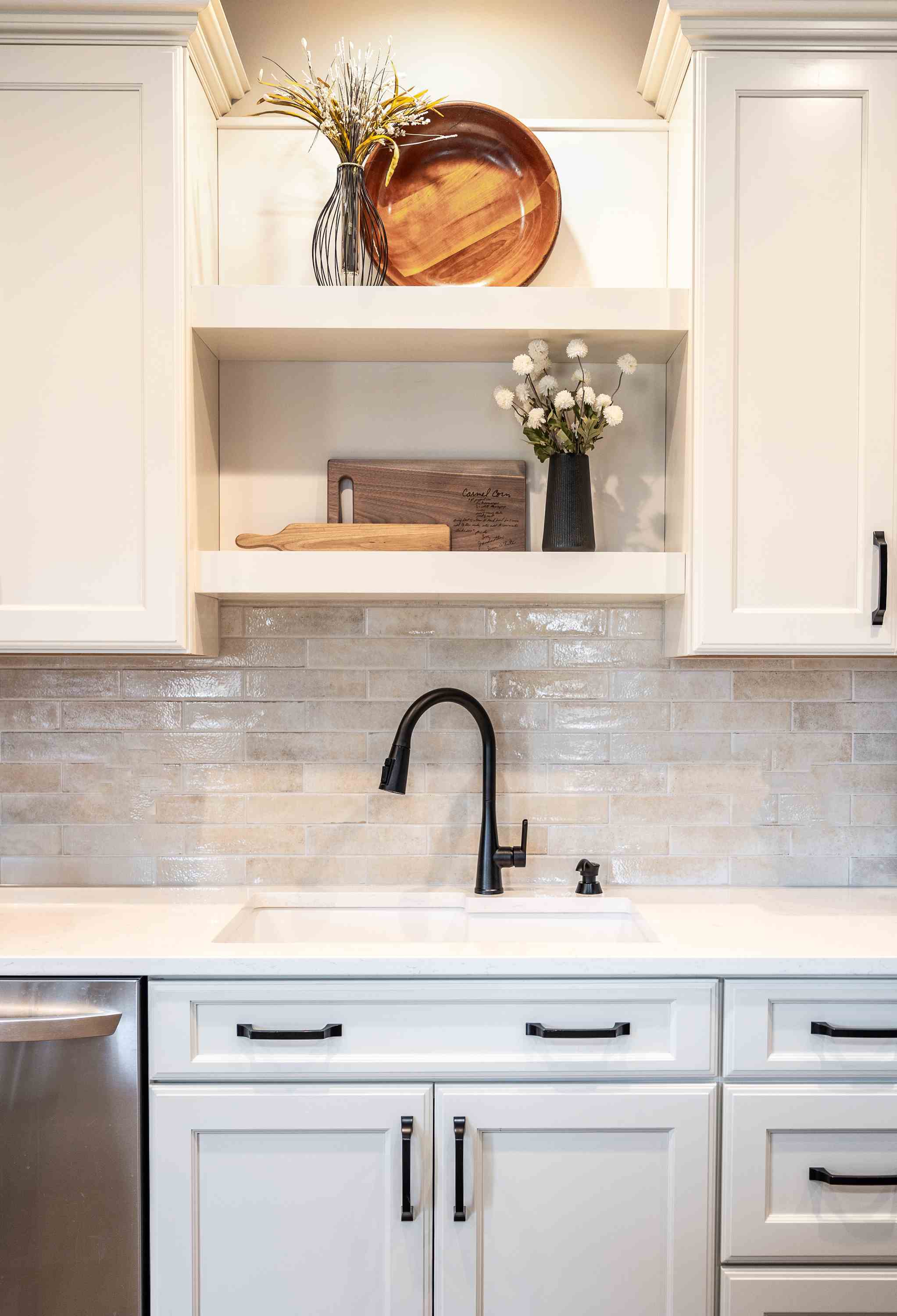 Modern kitchen sink with black faucet, white cabinets, beige tile backsplash, wooden cutting boards, and decorative vases with flowers.