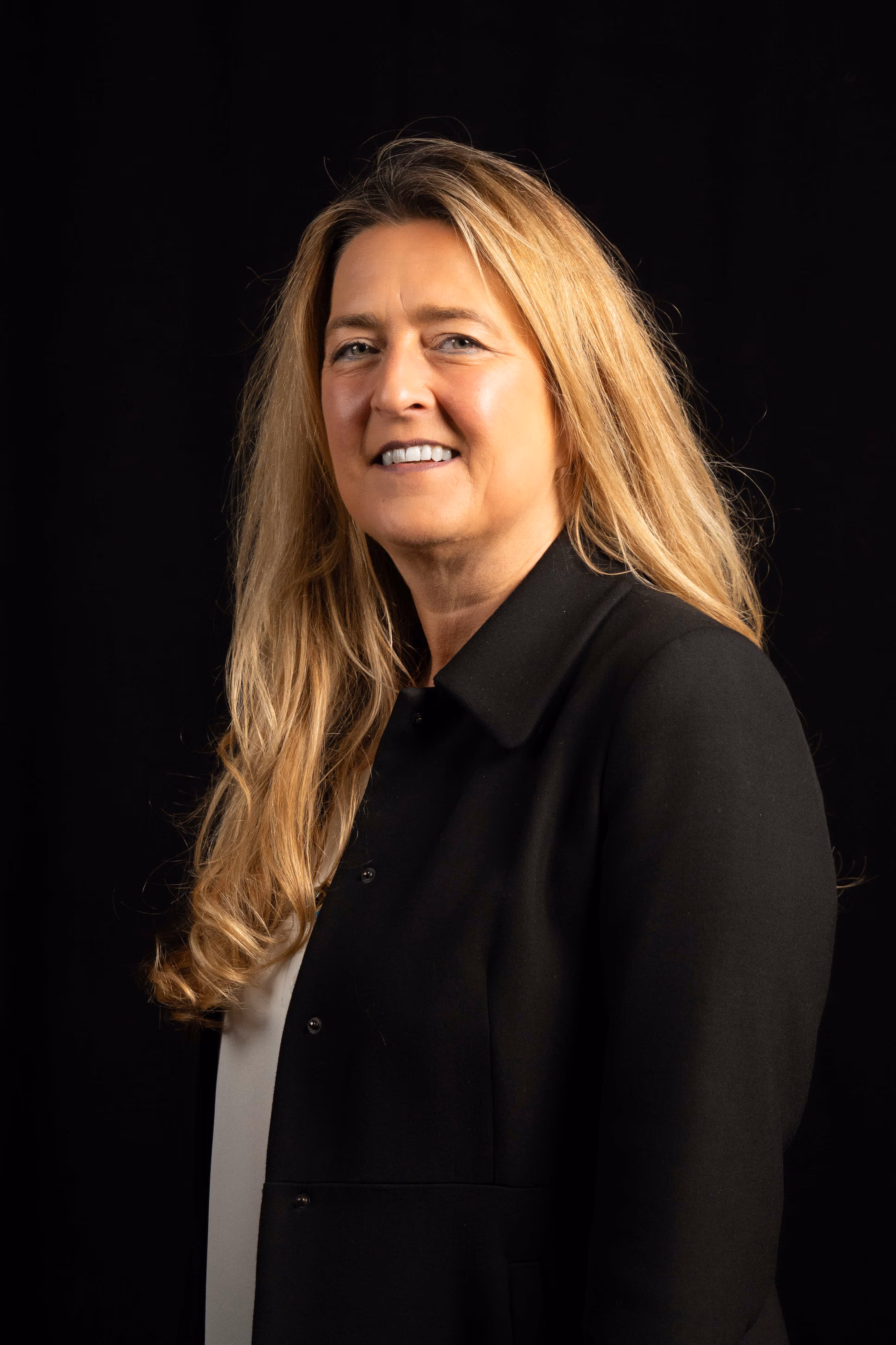 Smiling middle-aged woman with long blonde hair wearing a black jacket against a black background.