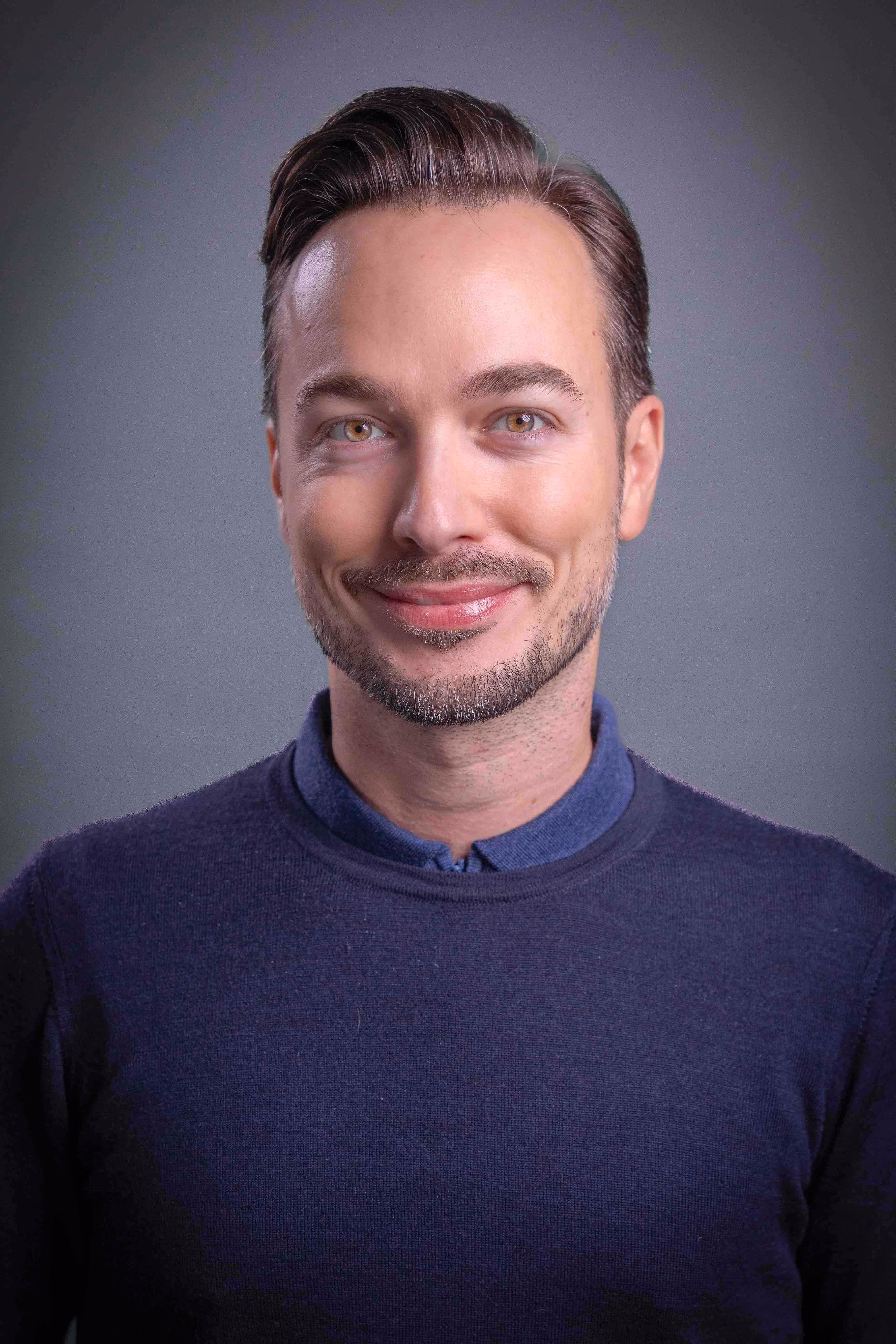 Smiling man with light stubble wearing a navy sweater over a blue collared shirt against a gray background.
