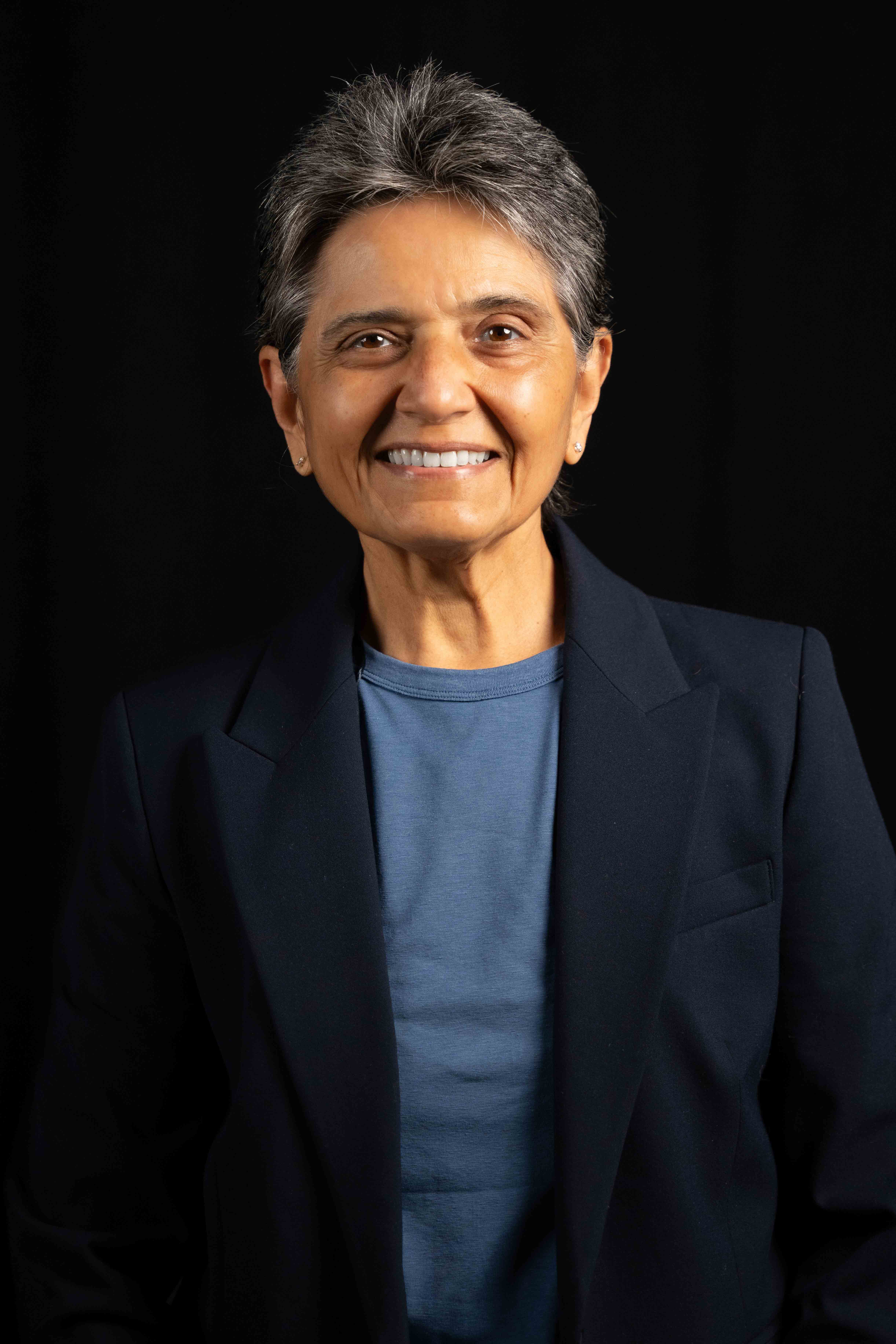 Smiling mature person with short gray hair wearing a blue shirt and dark blazer against a black background.