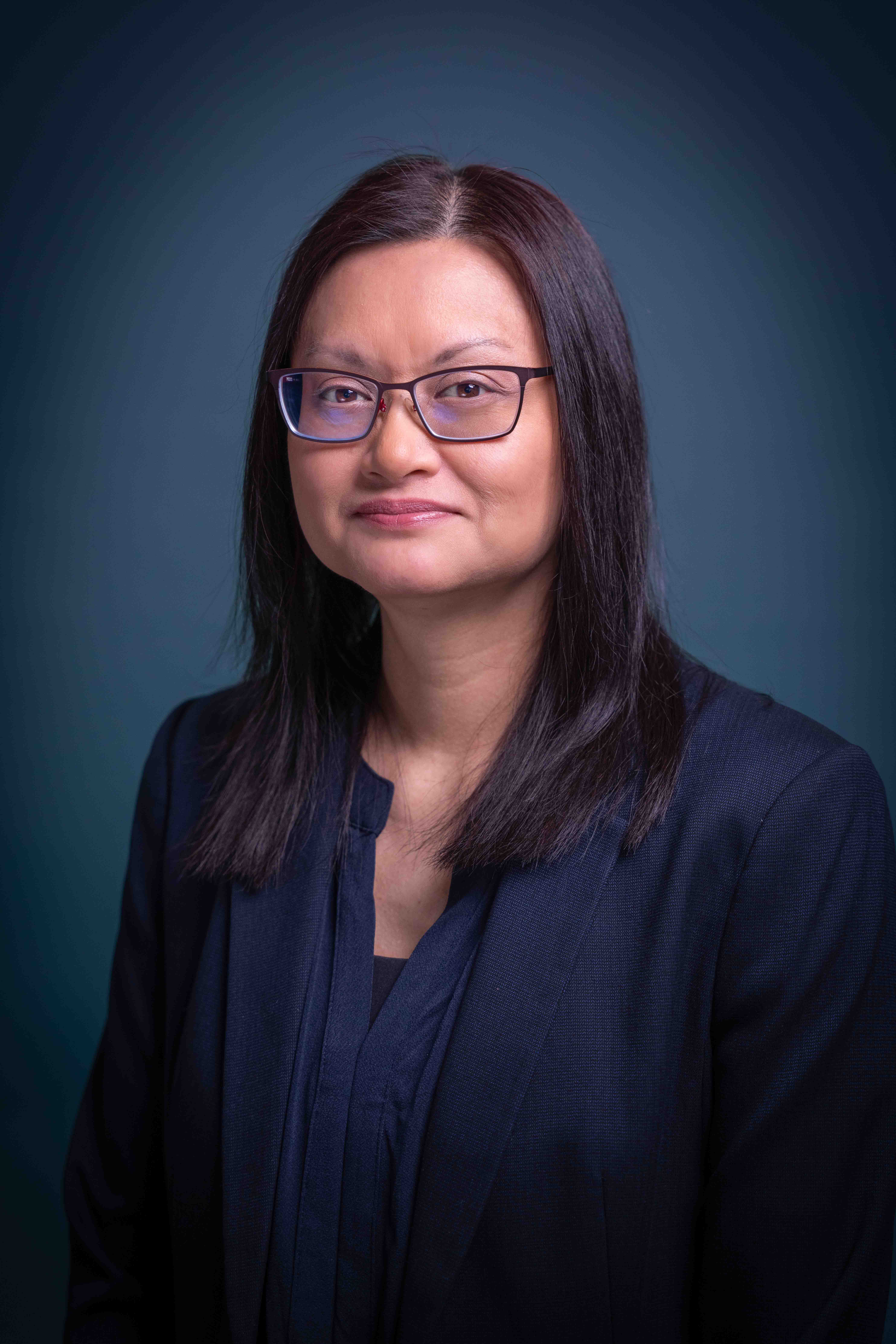 Professional woman with straight black hair and glasses wearing a dark blazer and blouse against a blue background.