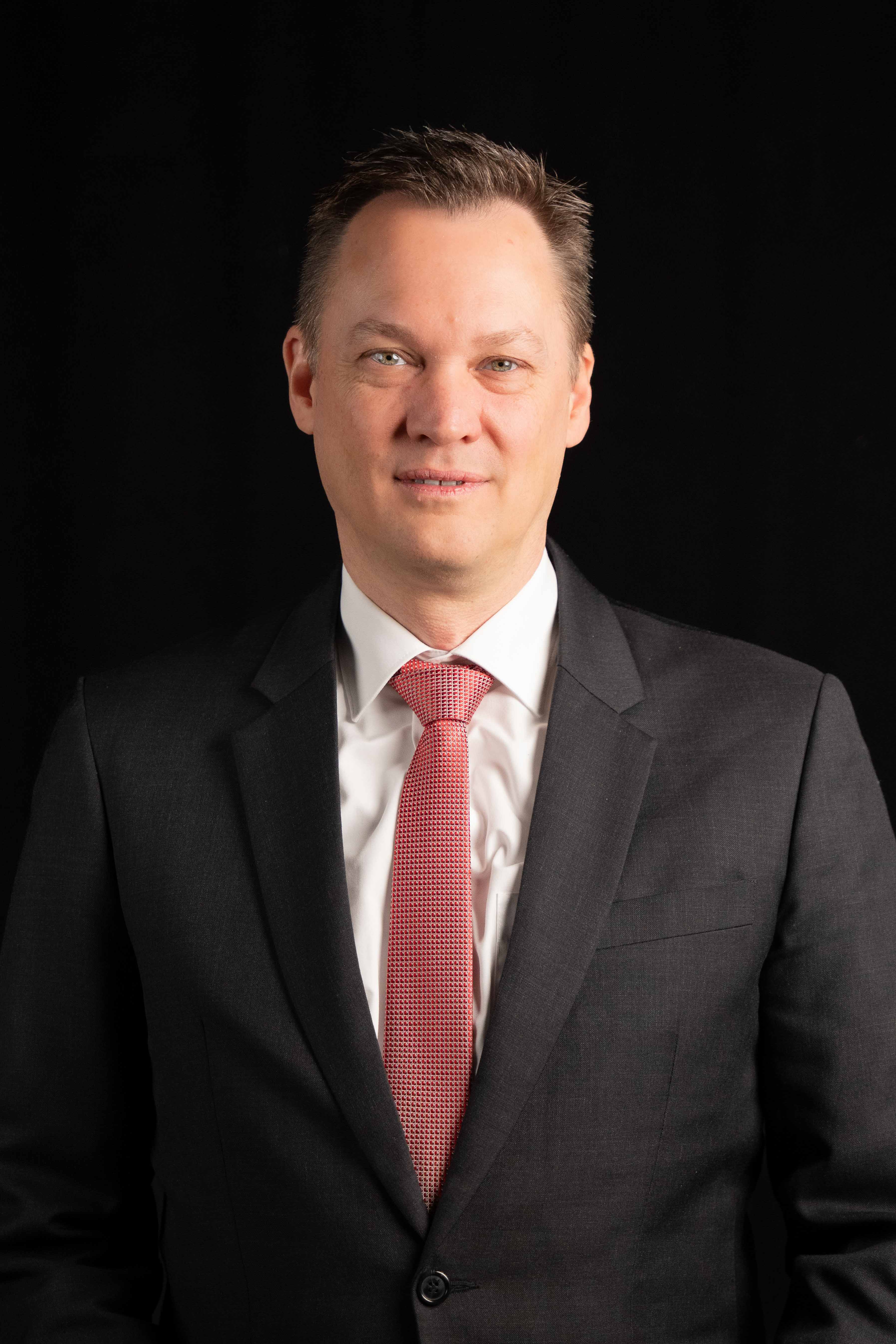 Smiling man wearing a black suit, white shirt, and red patterned tie against a black background.