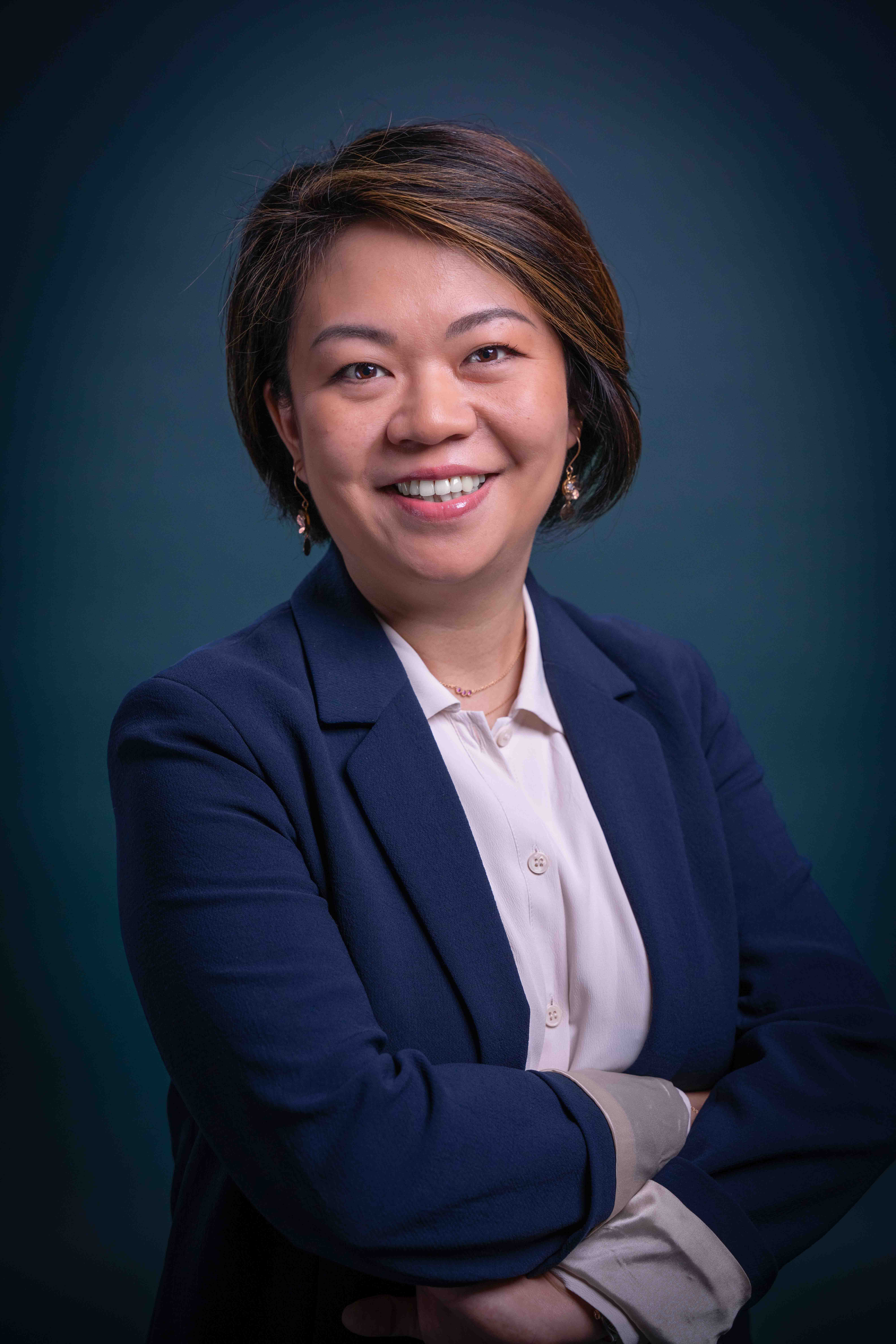 Smiling professional woman with short hair wearing a navy blazer and light blouse, standing with arms crossed against a dark background.