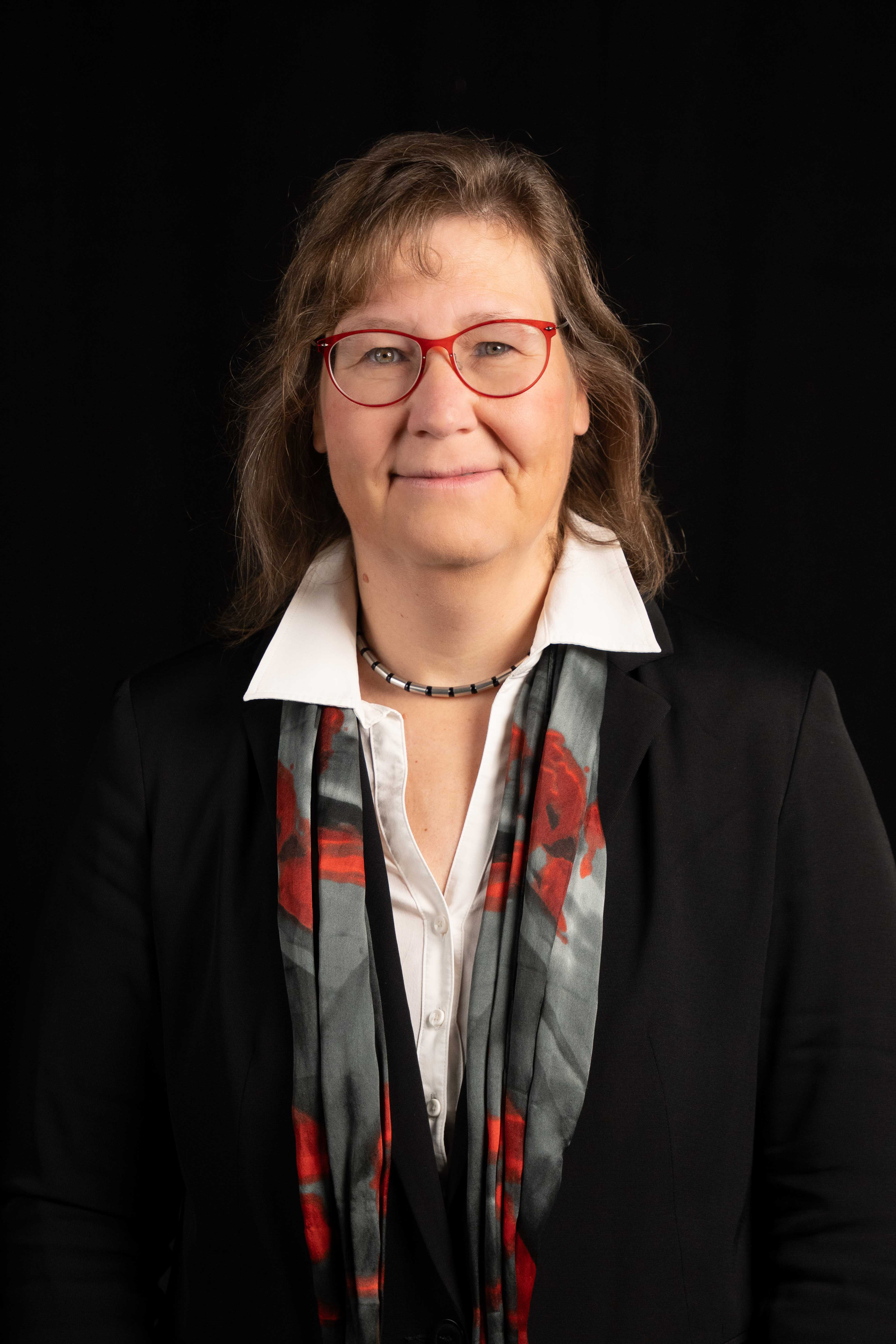 Smiling woman wearing red glasses, a white collared shirt, black blazer, and a gray scarf with red patterns, against a black background.