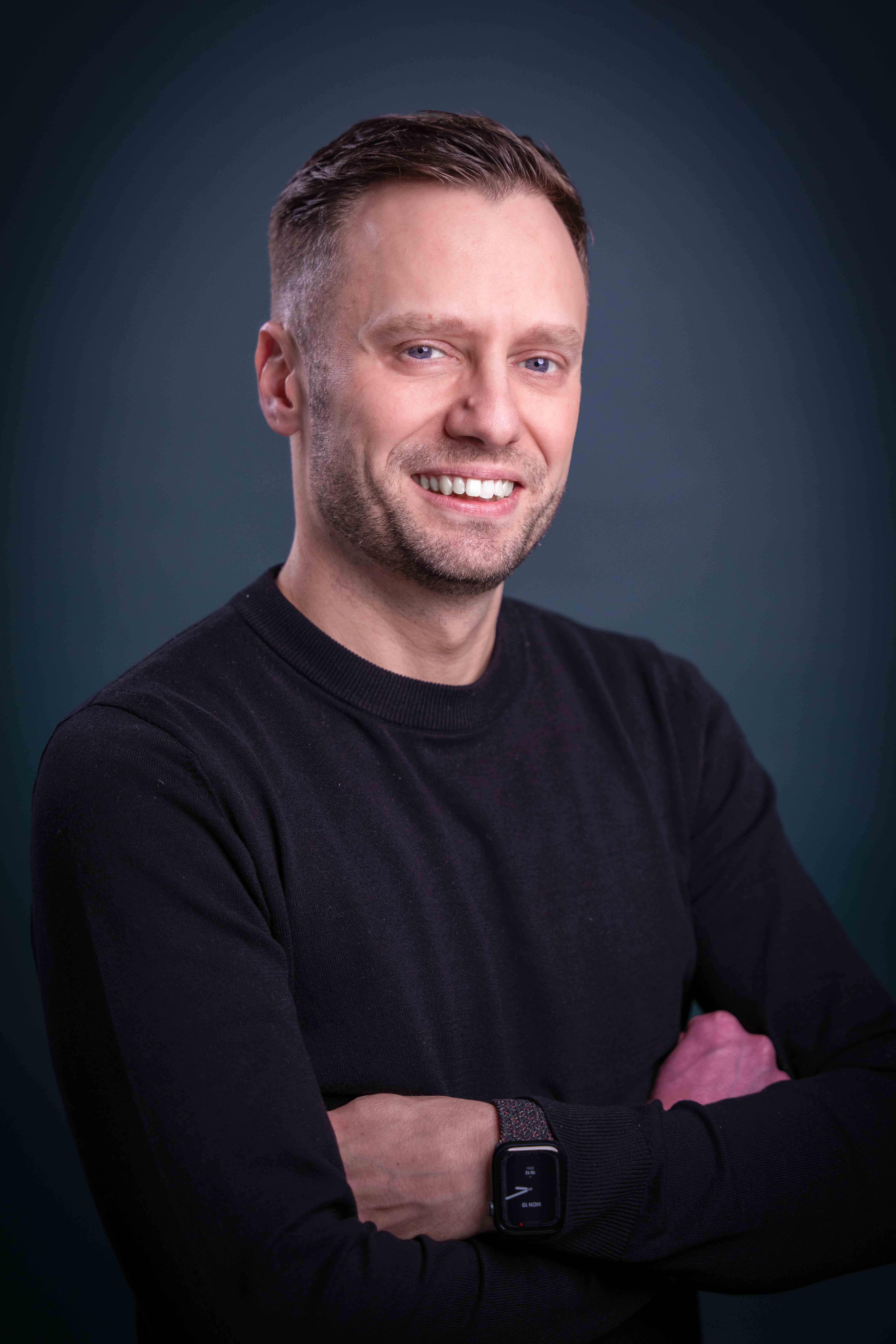 Smiling man with short hair wearing a black sweater and a smartwatch, standing against a dark blue background with arms crossed.