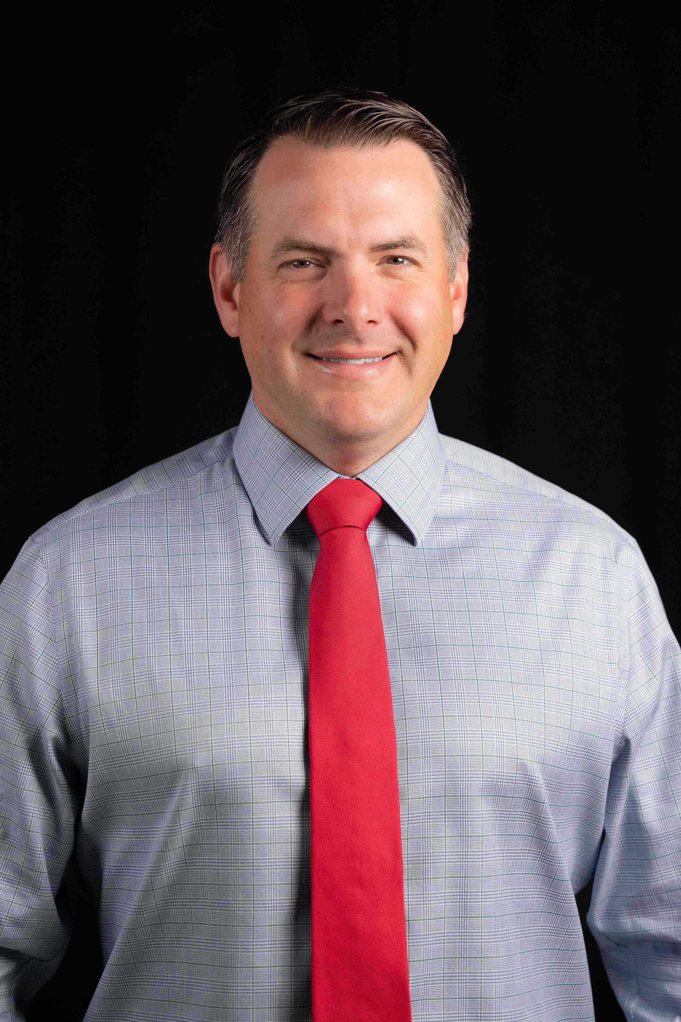 Smiling man wearing a grey checkered shirt and a red tie against a black background.