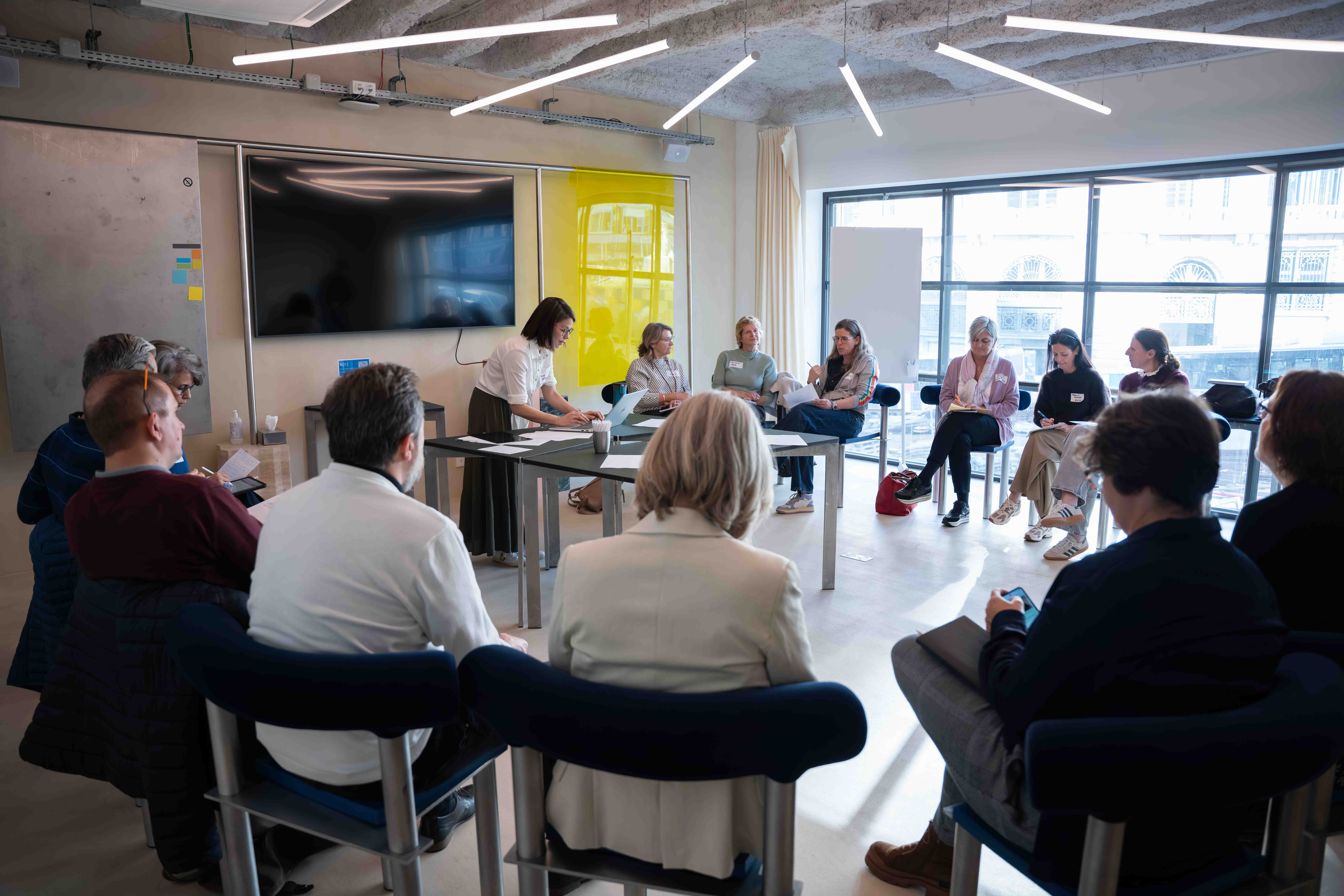 Group of people seated in a circle in a bright meeting room, one woman standing and typing on a laptop at a table.