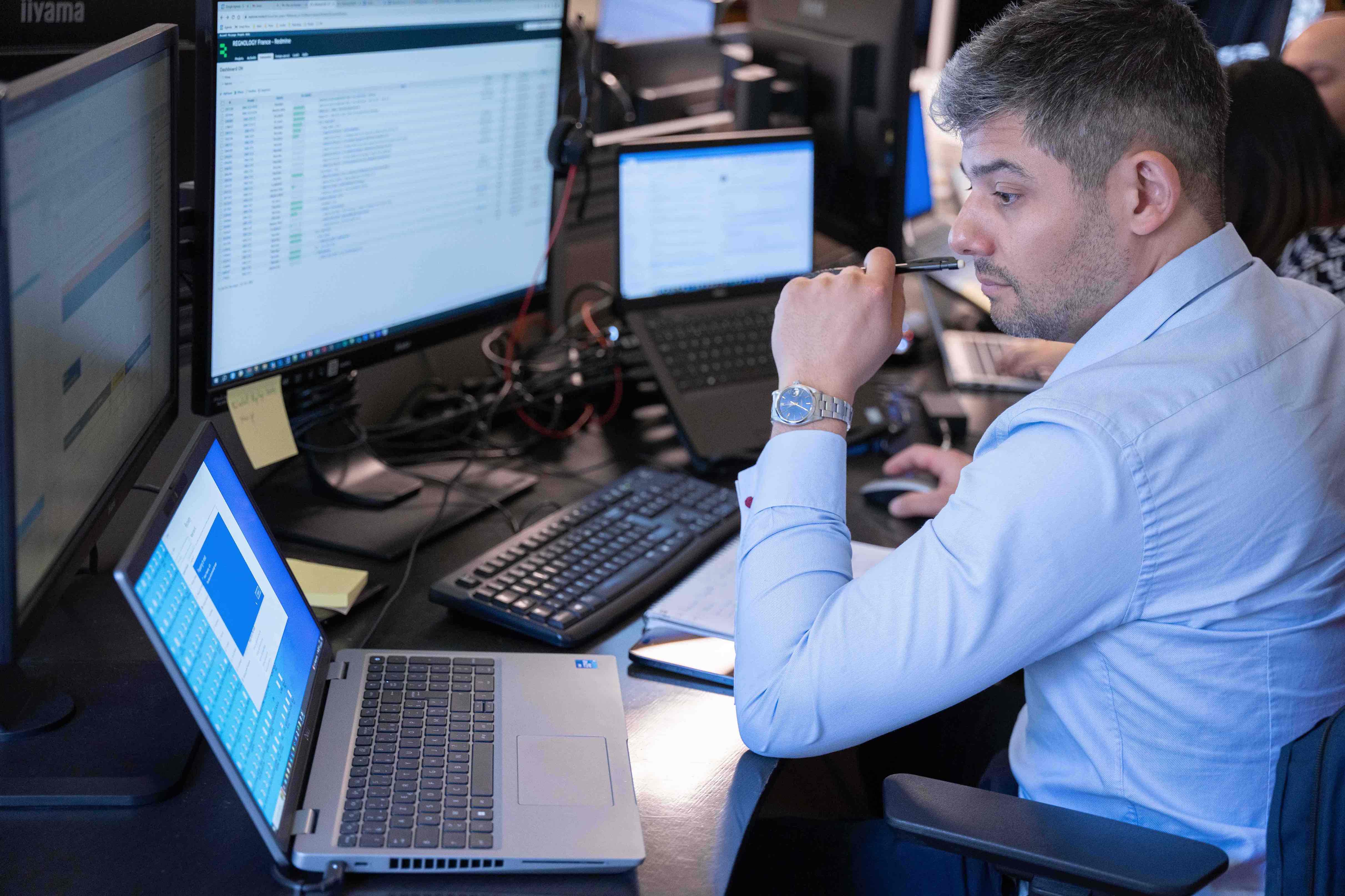 Man in a blue shirt sitting at a desk with multiple computer screens, holding a pen near his mouth and looking thoughtfully at the monitors.