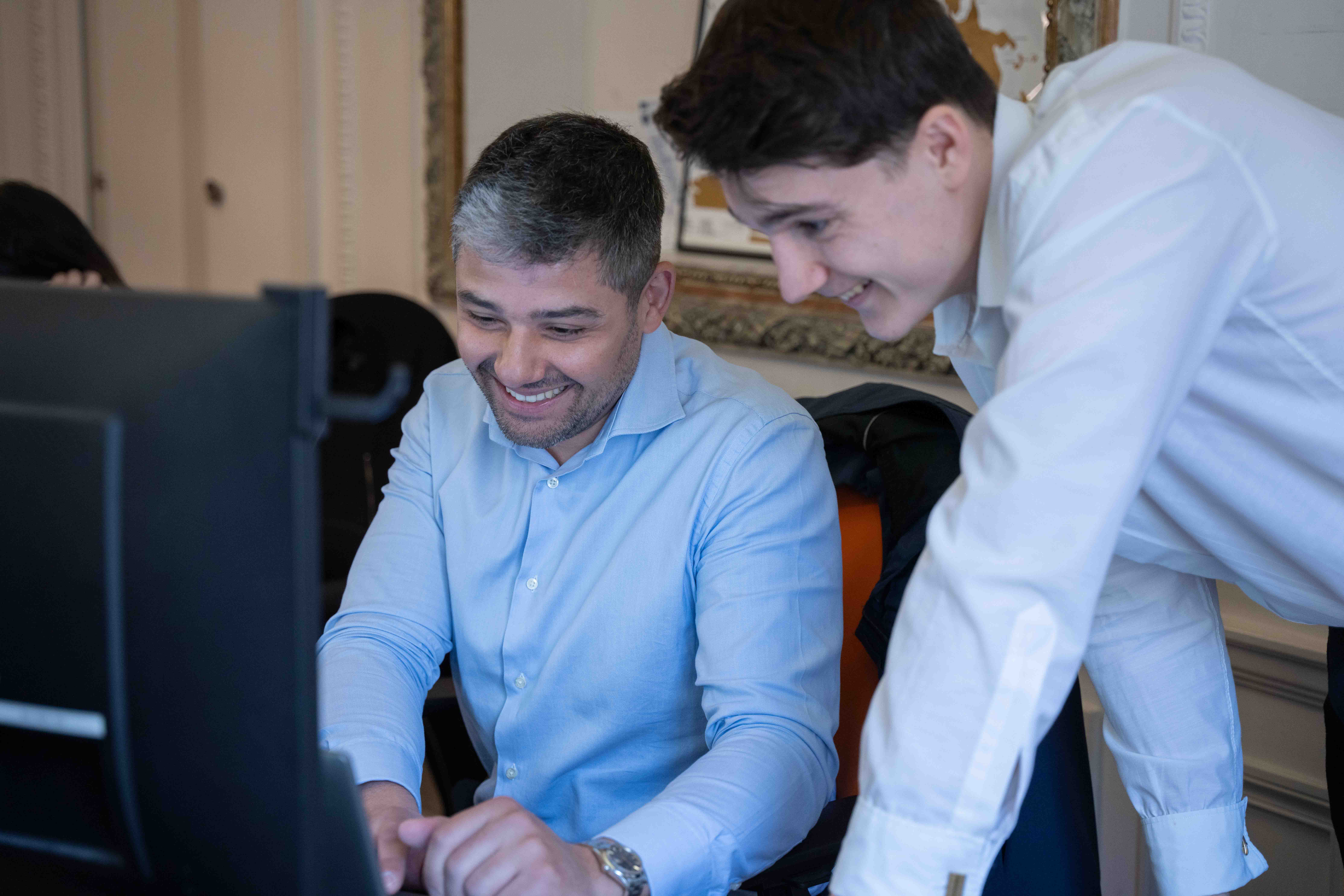 Two men smiling and working together at a computer in an office setting.