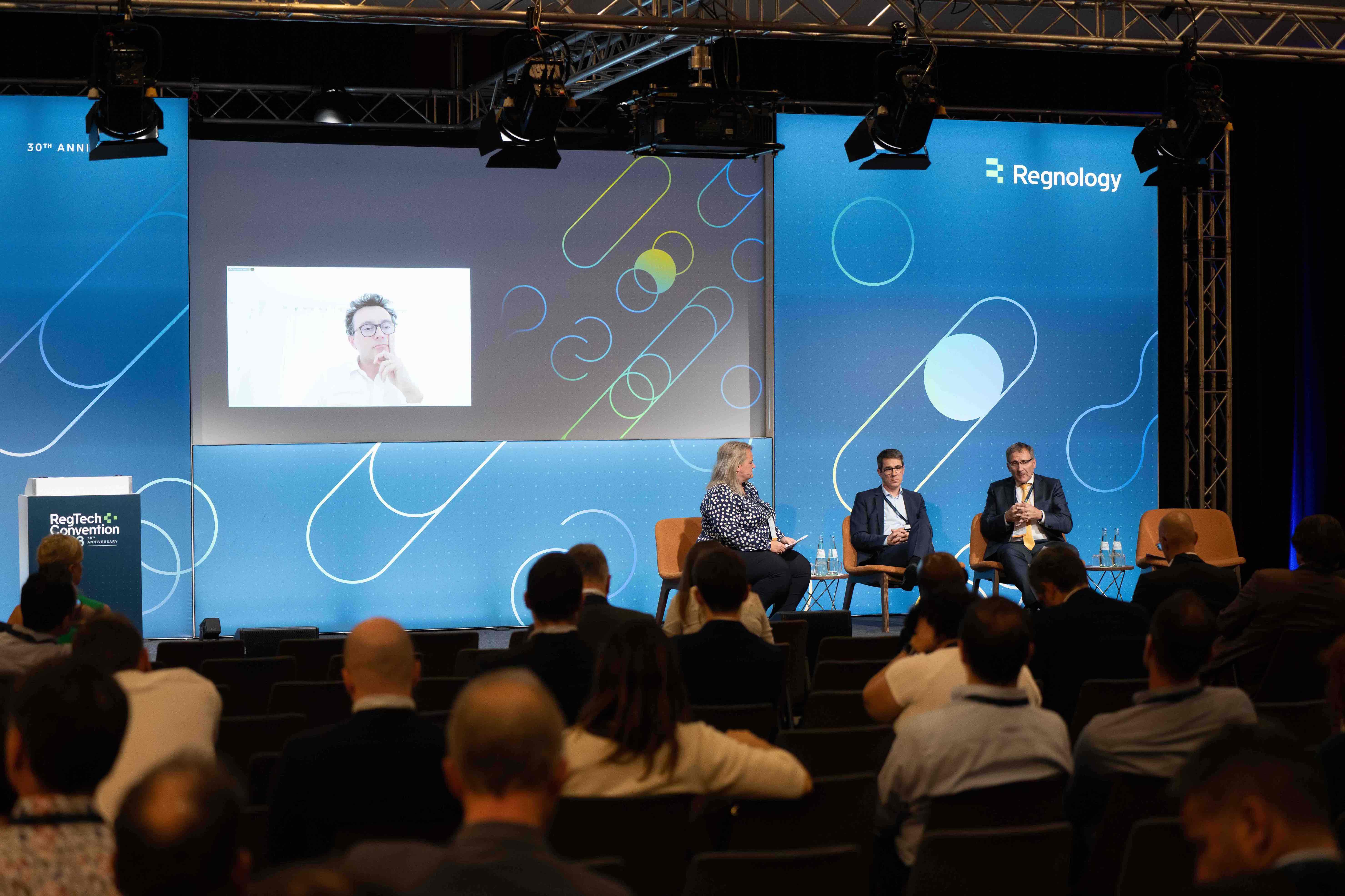 Panel discussion at RegTech Convention with three seated speakers on stage and one person participating via a video call projected on a screen.