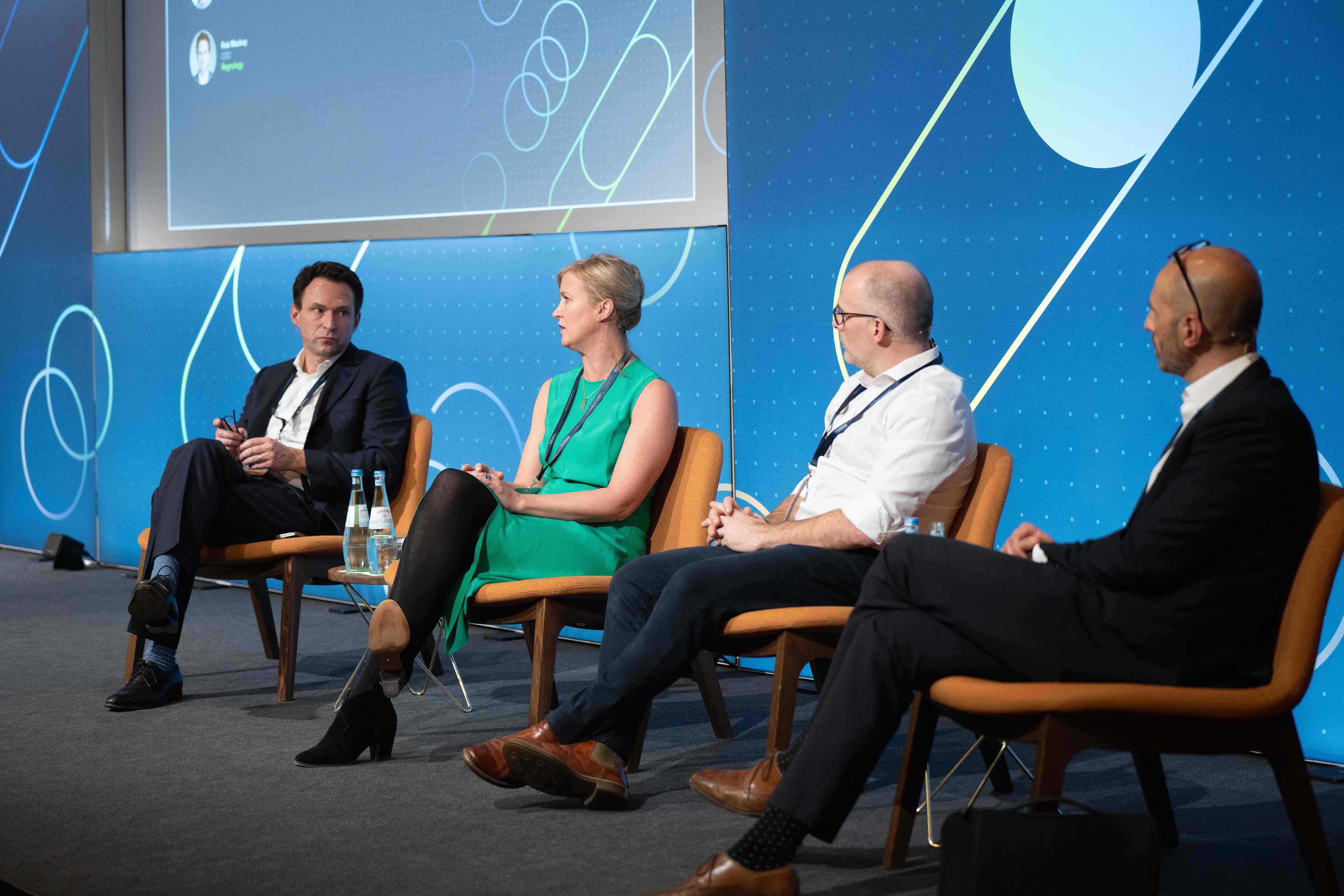 Four business professionals seated on stage in a panel discussion with blue abstract background.