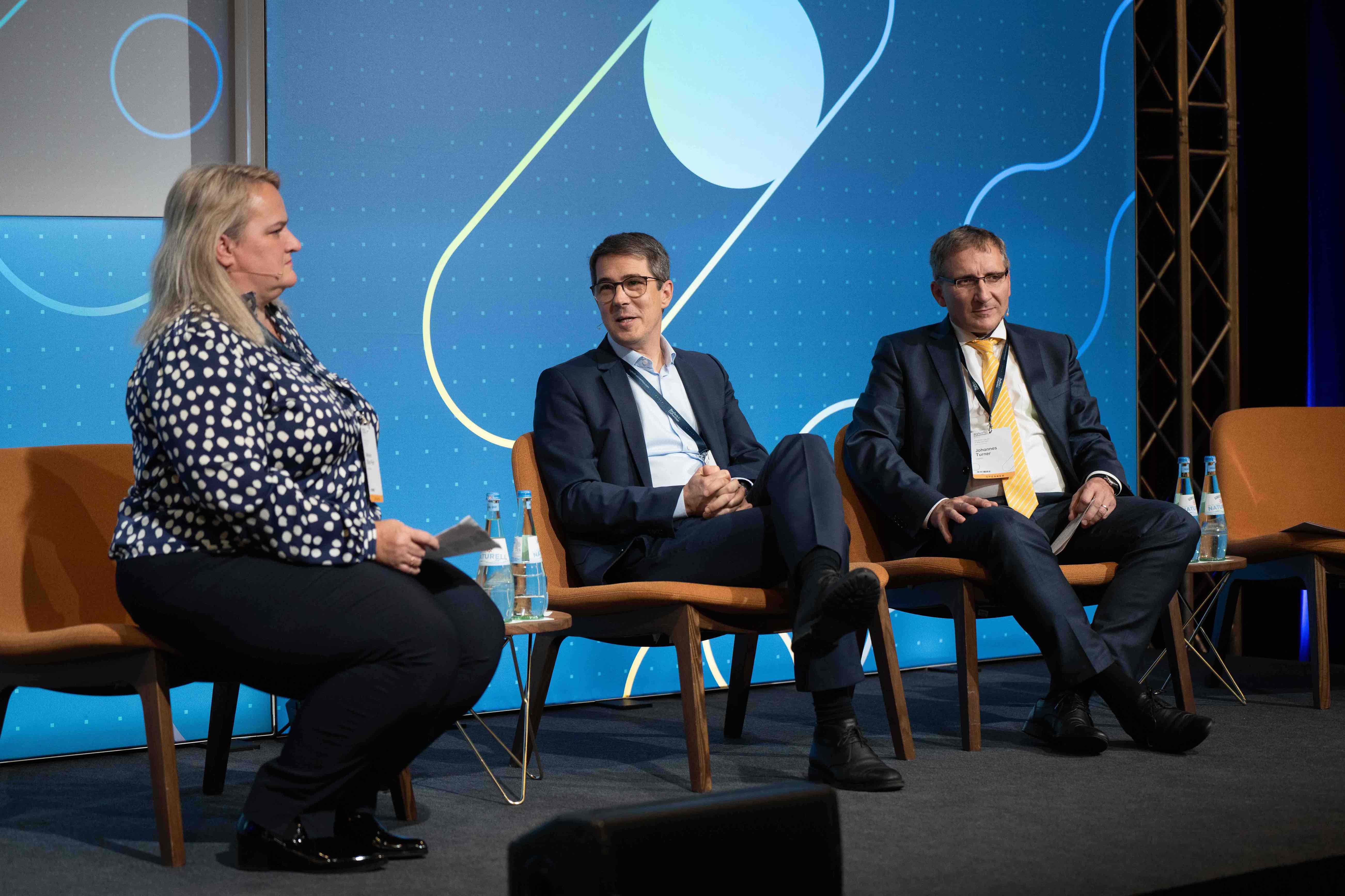 Three people seated on stage chairs during a panel discussion, with a blue abstract background and small tables holding bottles of water.