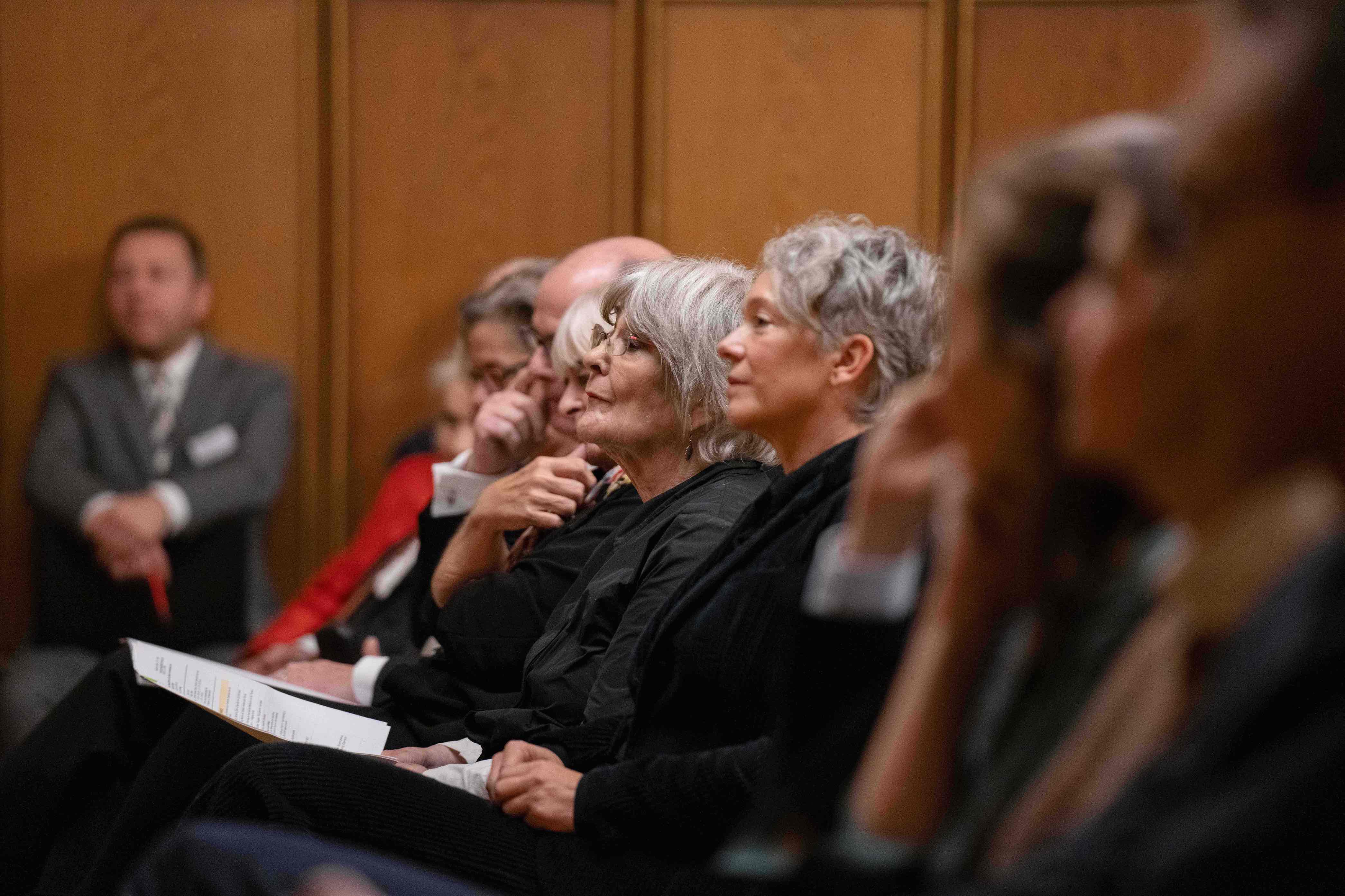Row of people seated attentively, including two older women with gray hair, in a wood-paneled room.
