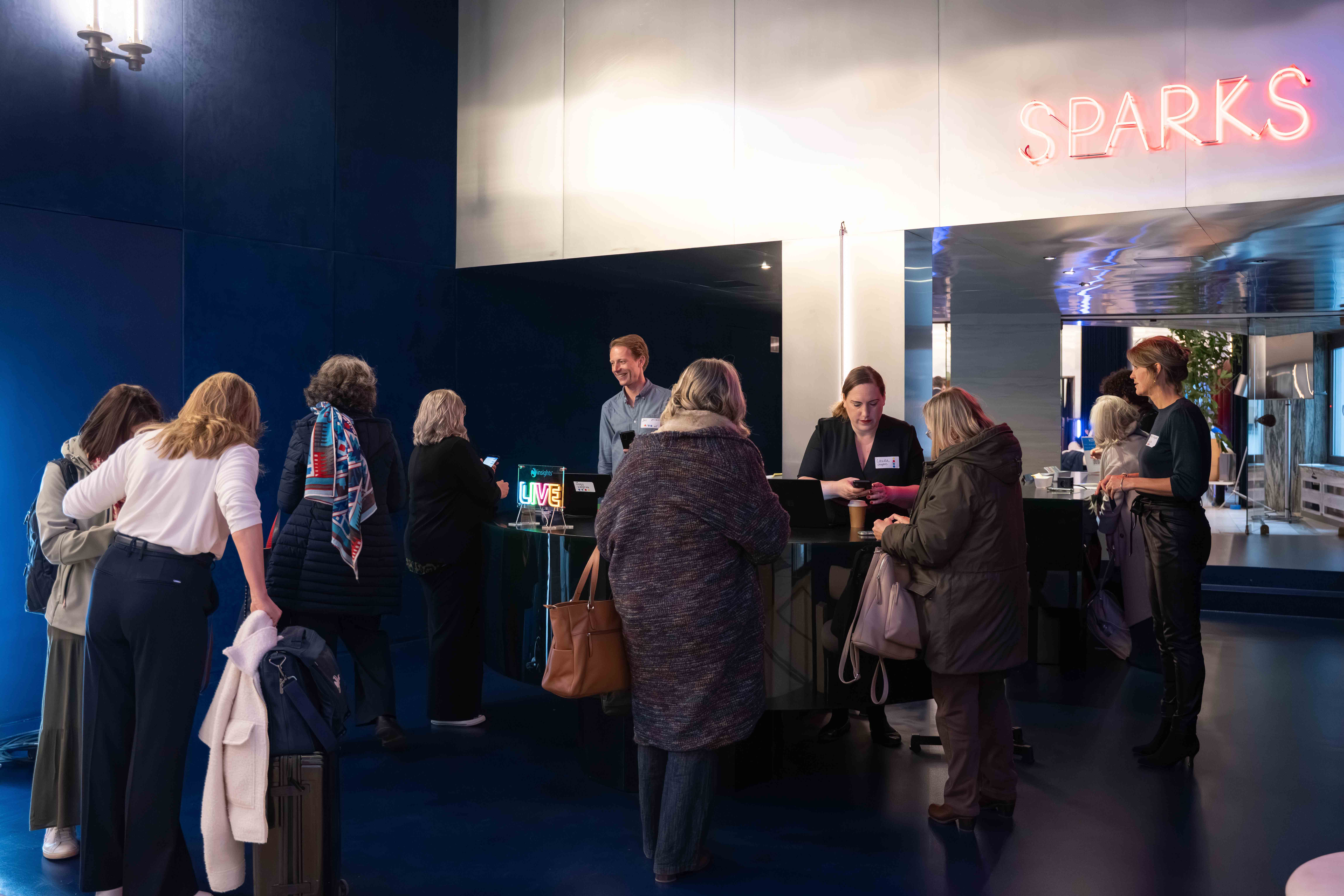 People lined up at a reception desk with staff checking them in under a neon sign that reads SPARKS.