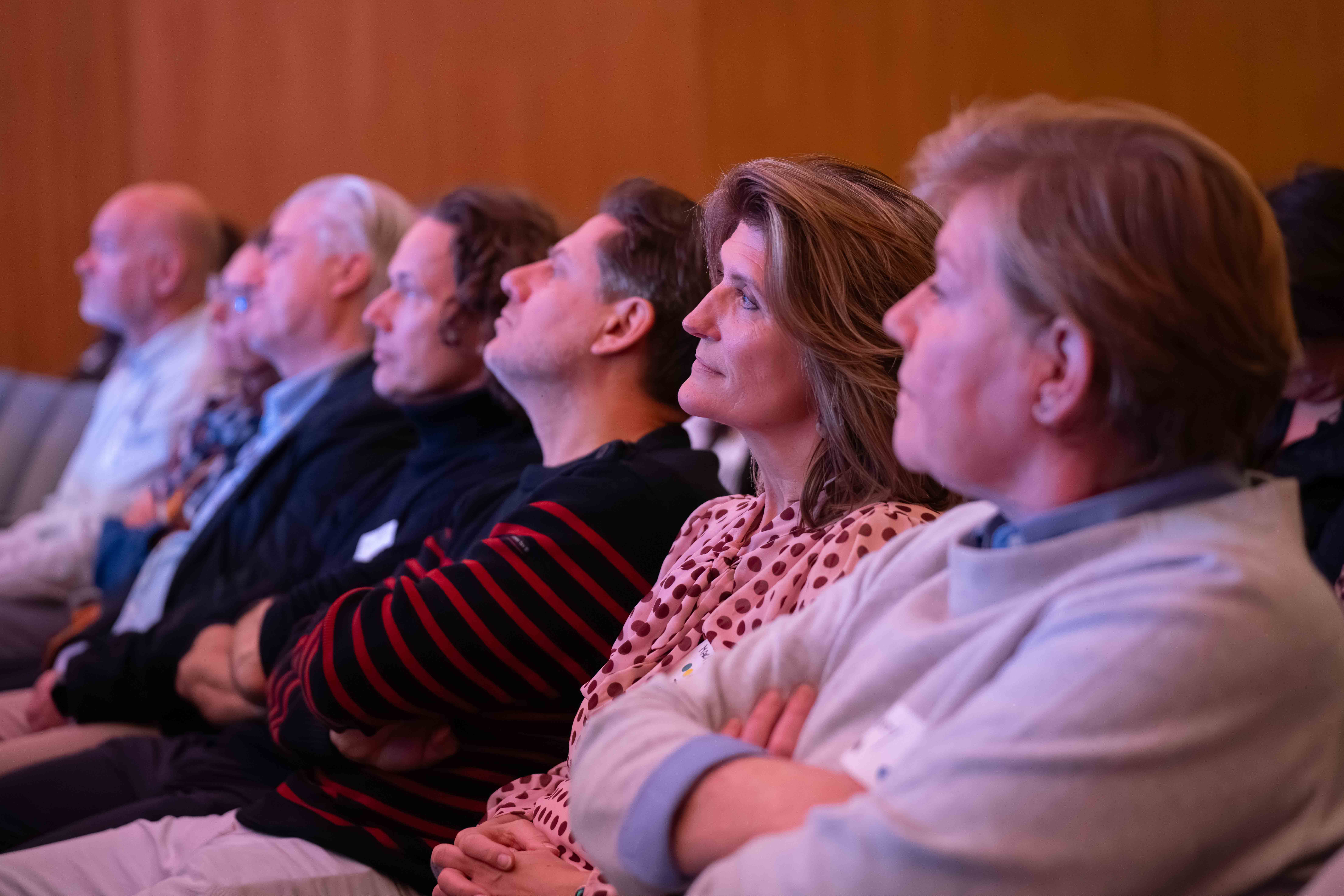 Row of adults seated indoors attentively watching or listening to a presentation.