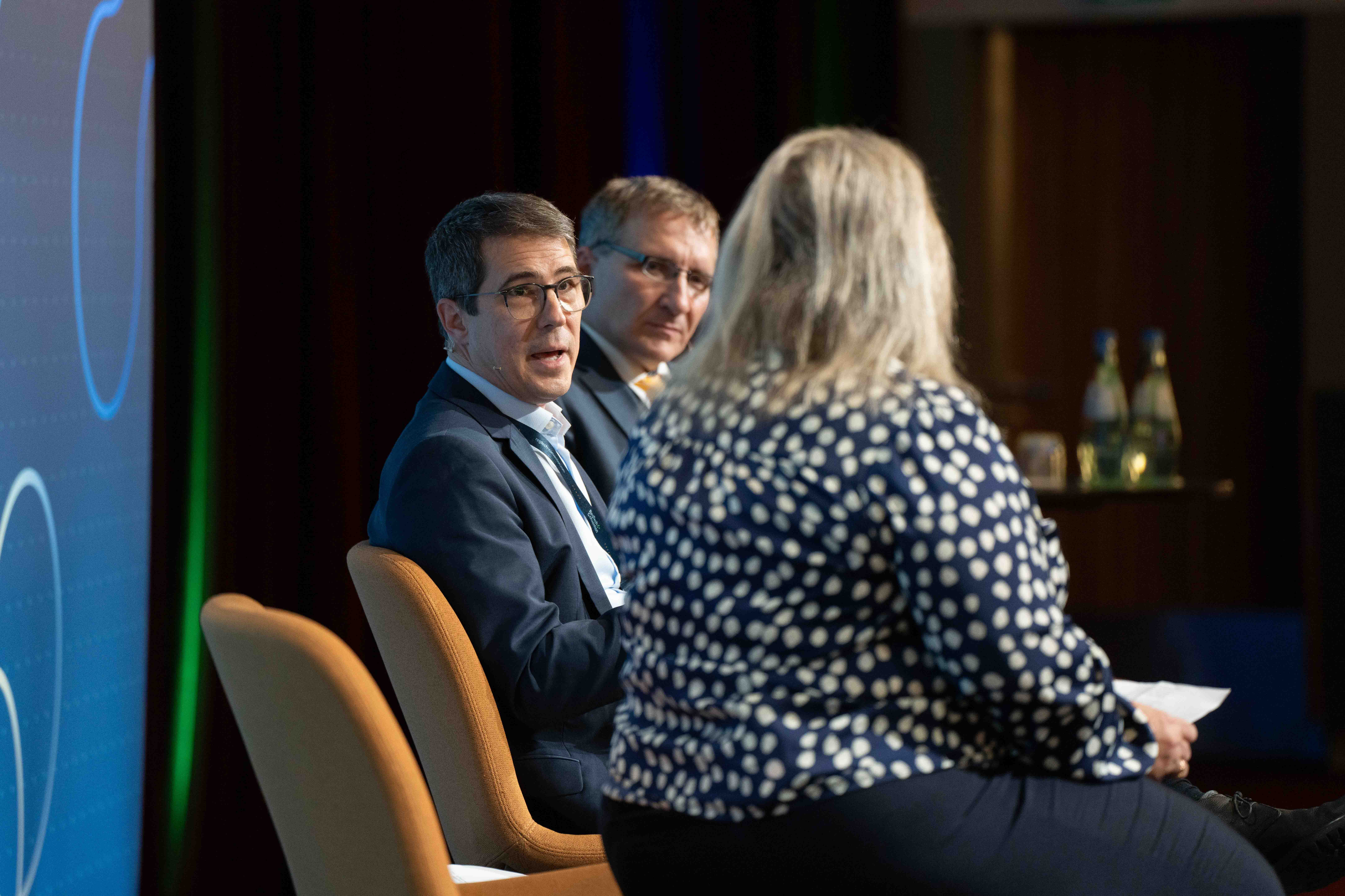 Two men in suits and a woman in a polka dot blouse engaged in a panel discussion on stage.
