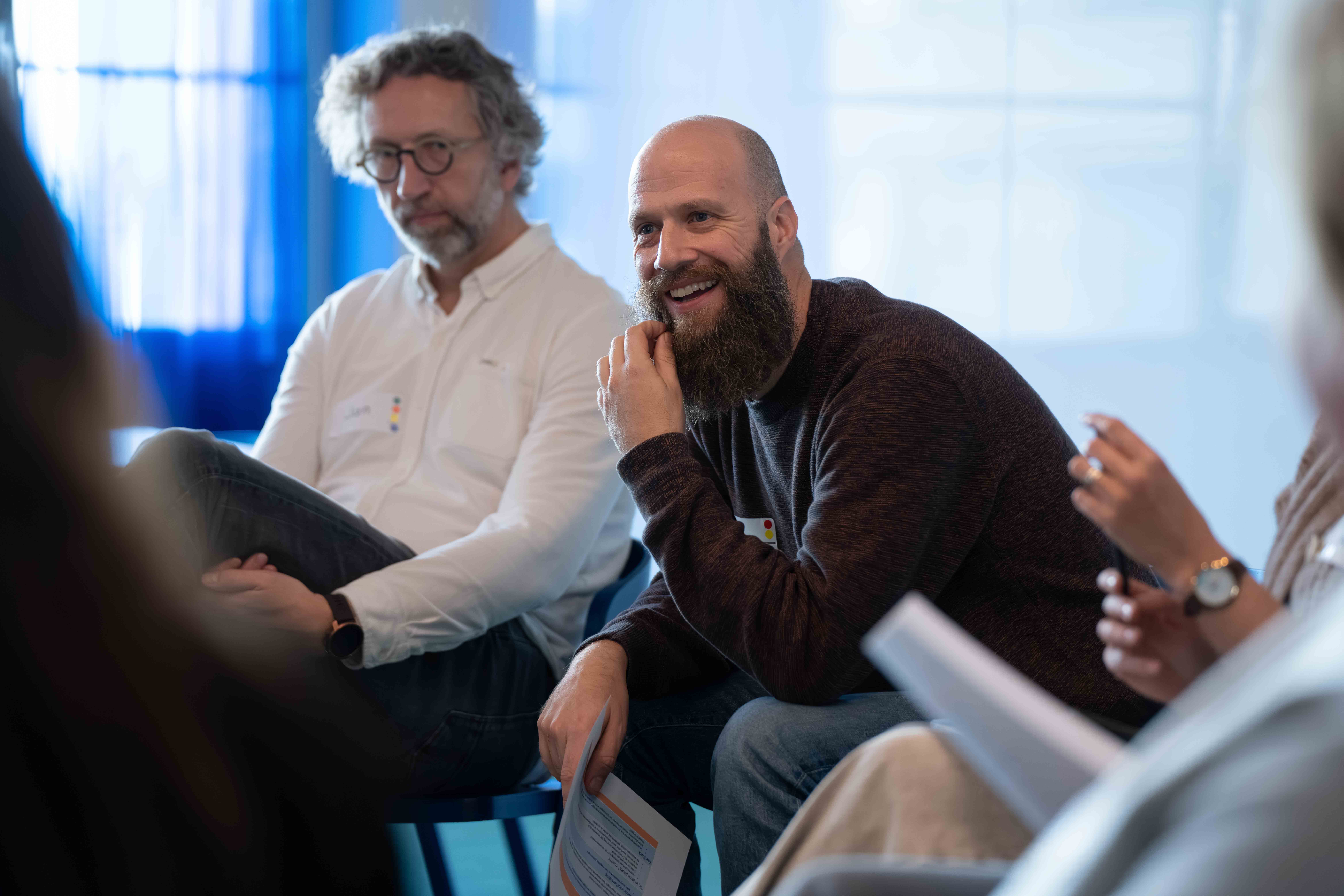 Two men seated indoors, one with glasses and white shirt, the other with a beard and dark sweater, engaged in a group discussion.