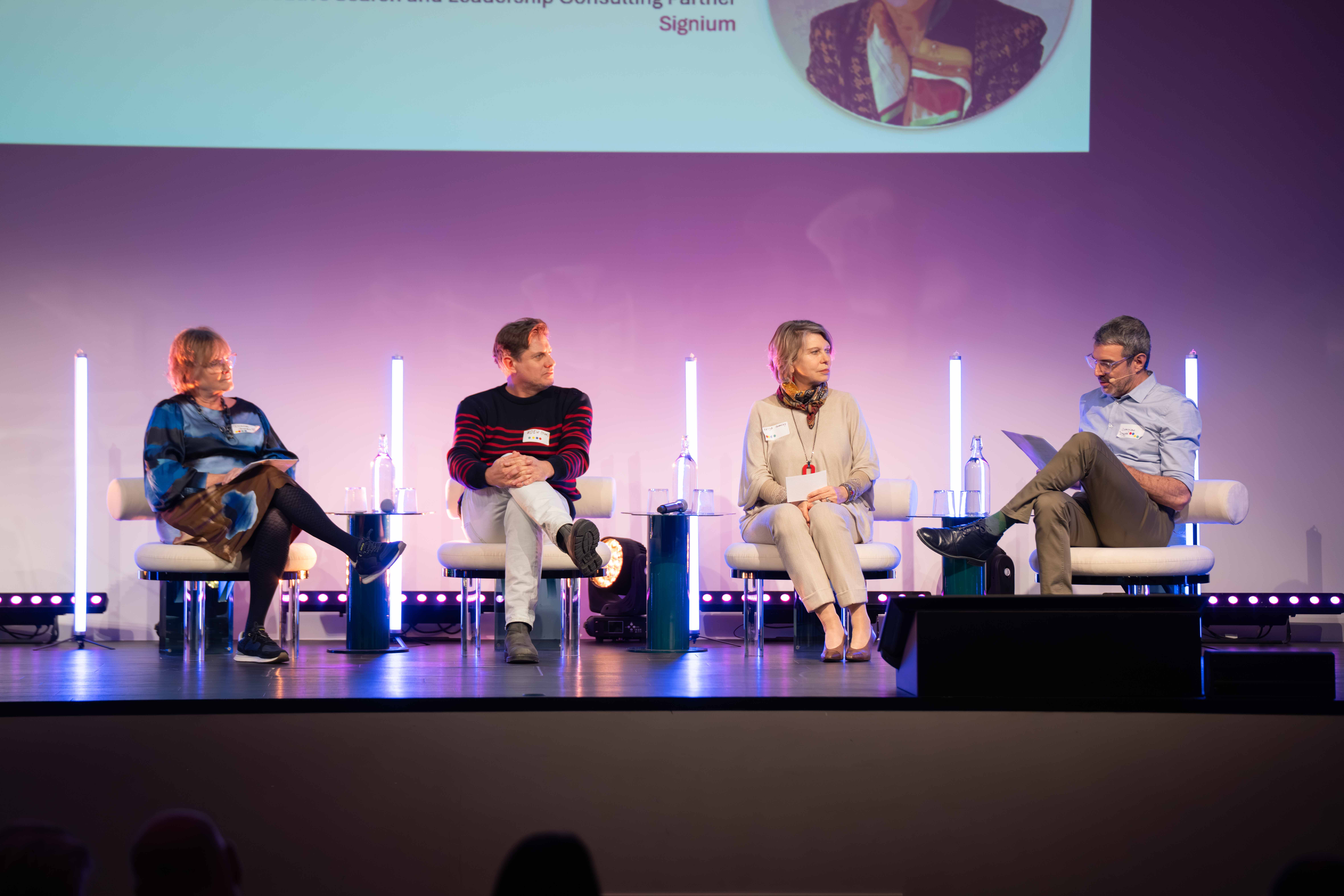 Four panelists seated on stage during a discussion, each with a microphone and water bottles between them.