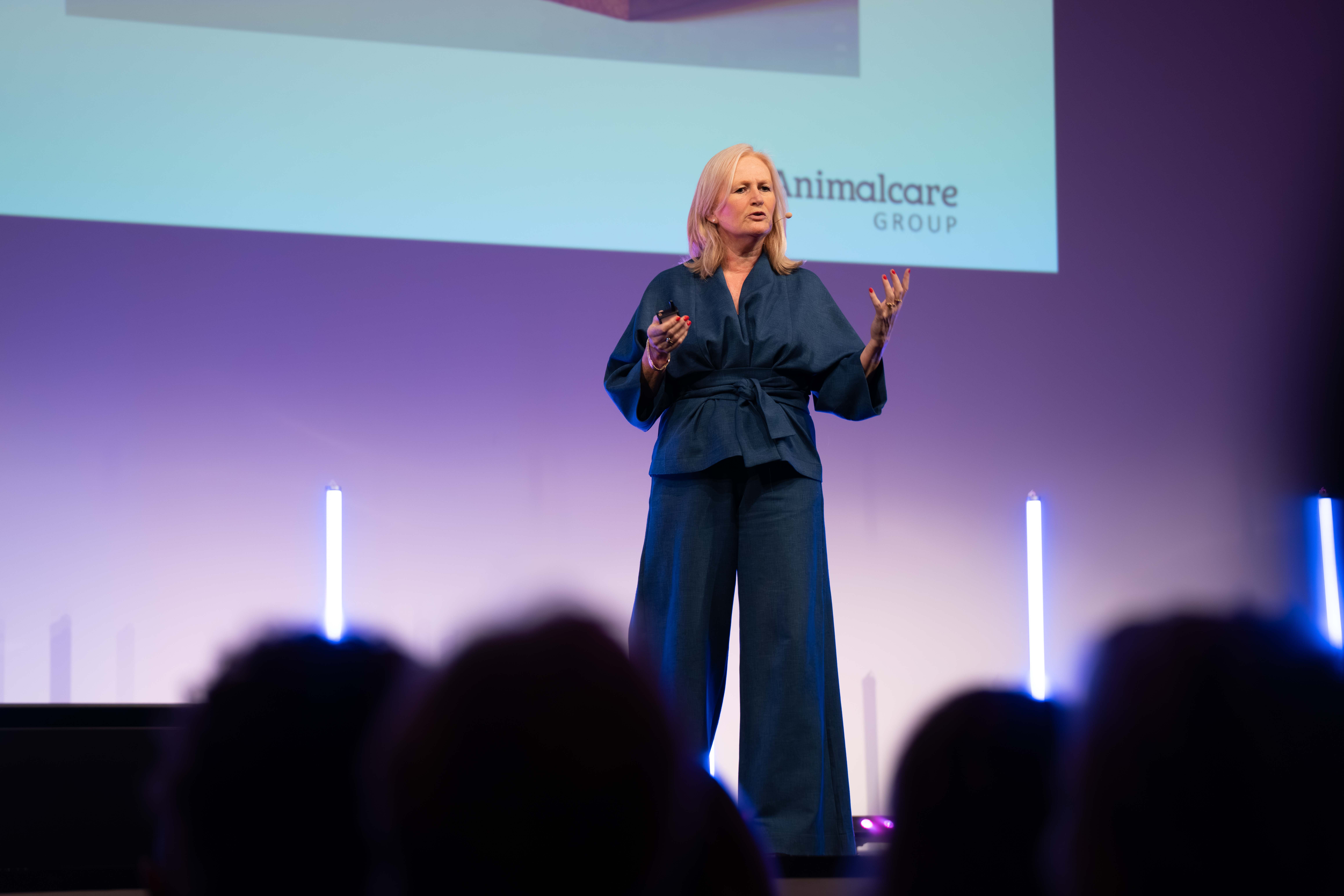 A woman in a blue outfit speaking on stage with a microphone headset and presentation slide behind her.