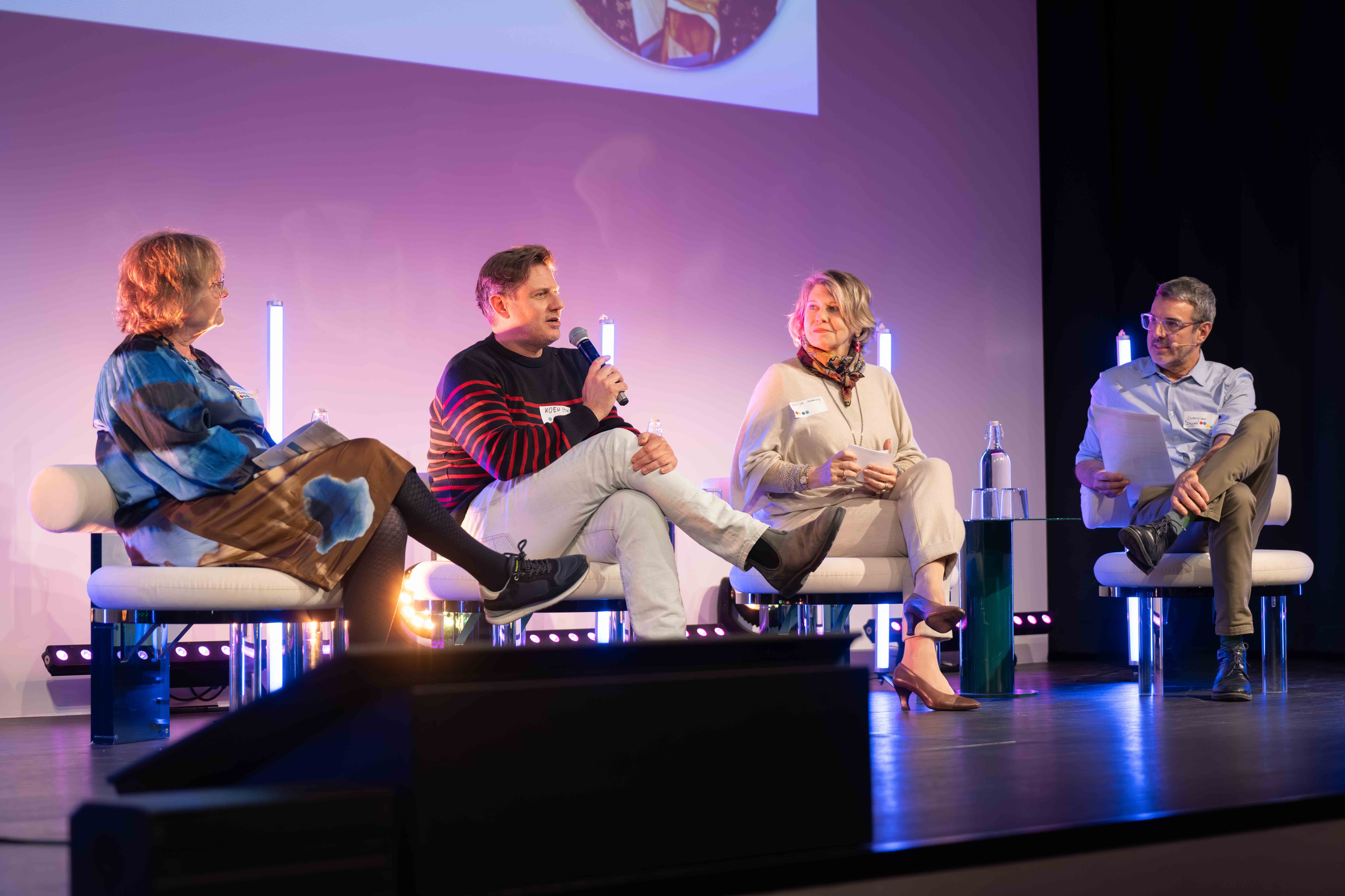 Four people seated on stage engaged in a panel discussion, one man is speaking into a microphone while others listen.